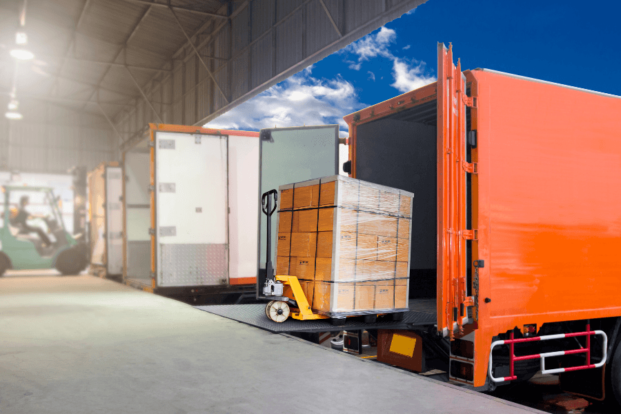 Forklift operator loading a shrink-wrapped wooden crate into a transport truck at a logistics facility.