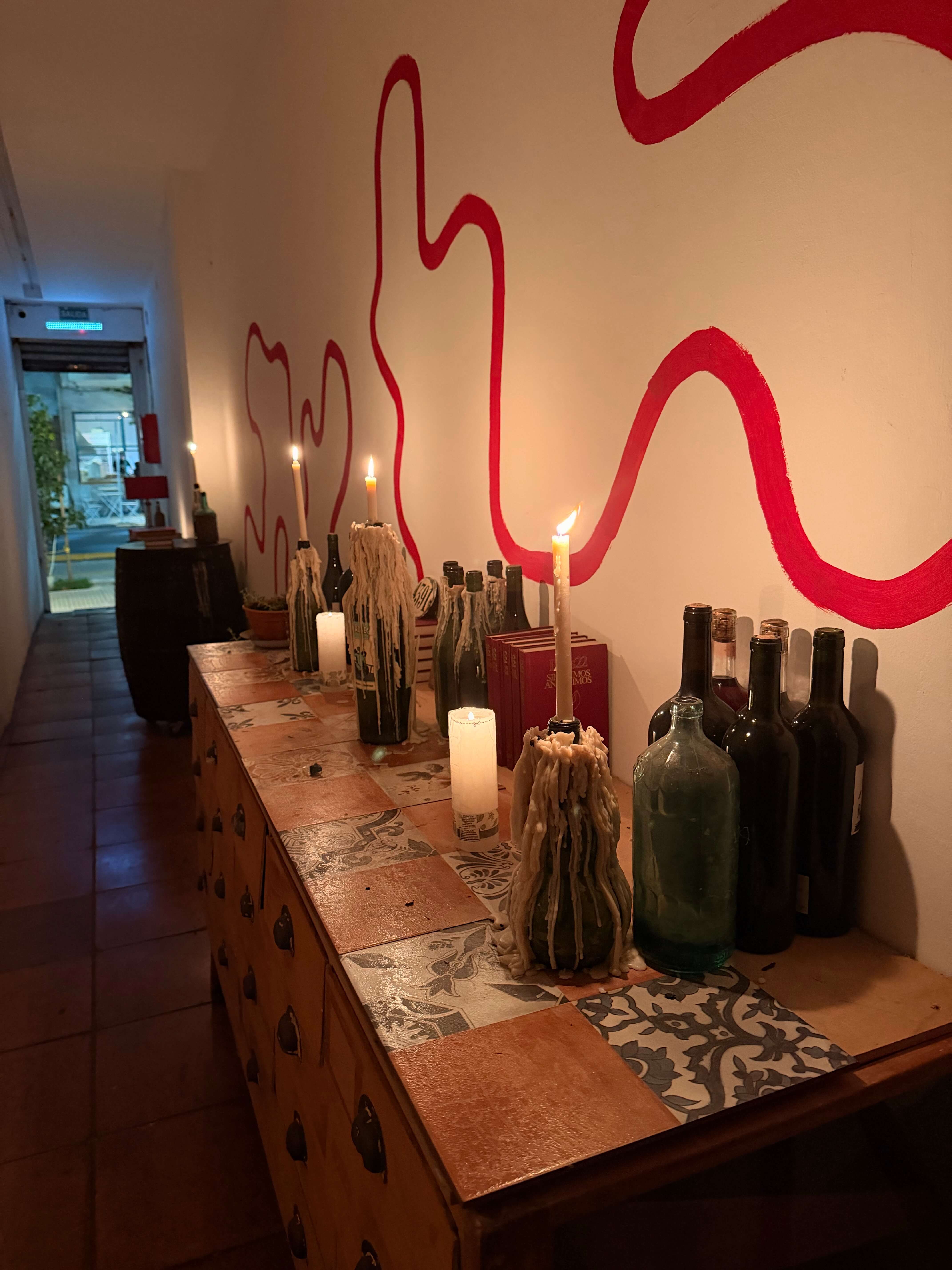 A table full of wine bottles and candlabras at dusk set against a wall with red squiggly lines painted across it