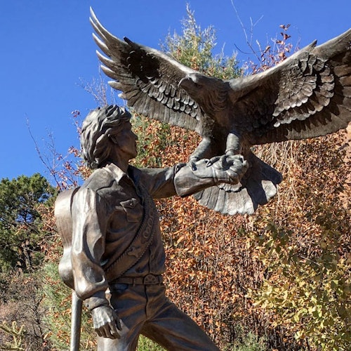 Bronze statue of a person holding a perched eagle, with trees and a clear blue sky in the background.