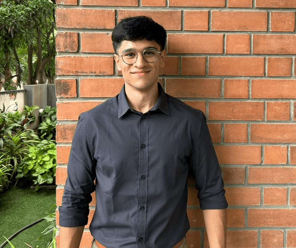 A person with medium-length brown hair wearing a dark, textured coat and a burgundy turtleneck. They have a calm, neutral expression.