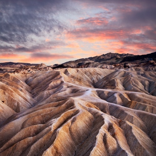 Layered, undulating rock formations under a dramatic sunset sky with pink and orange hues. No people are present.