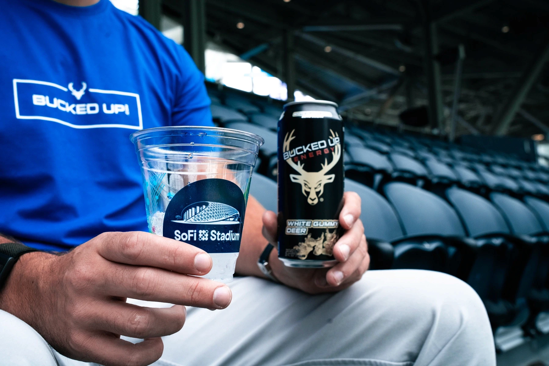 A person seated at SoFi Stadium holds a Bucked Up Energy White Gummy Deer can in one hand and a stadium-branded plastic cup in the other, wearing a bright blue “Bucked Up” t-shirt. The focus highlights the energy drink branding and casual game-day vibe.