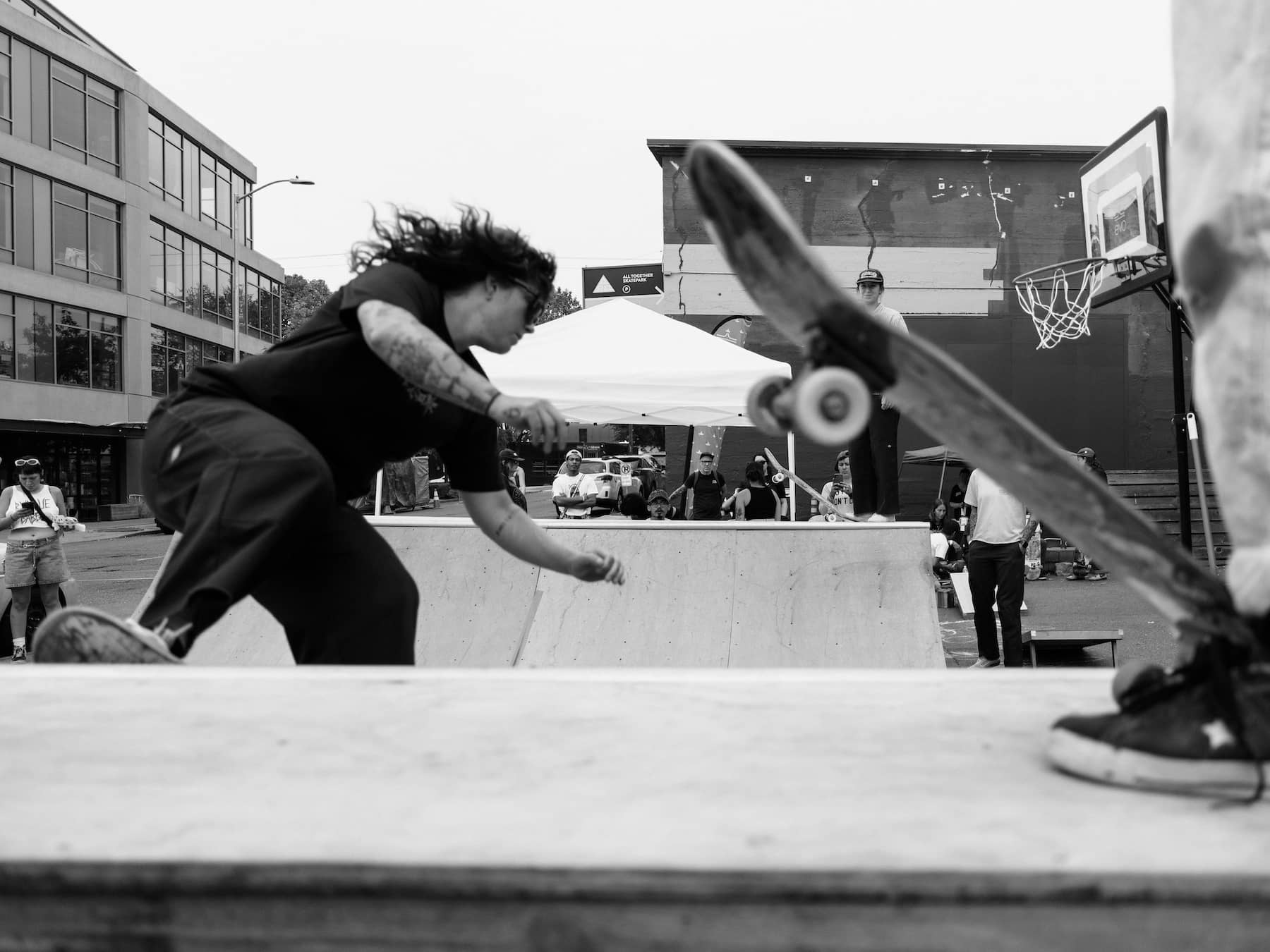 Person skateboarding in mid-air during a street skateboarding event, with onlookers and a basketball hoop in the background.