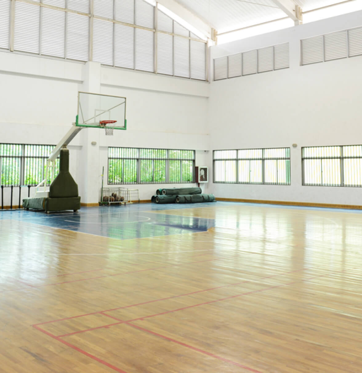 A bright indoor basketball court with a hoop, large windows, and a polished wooden floor.