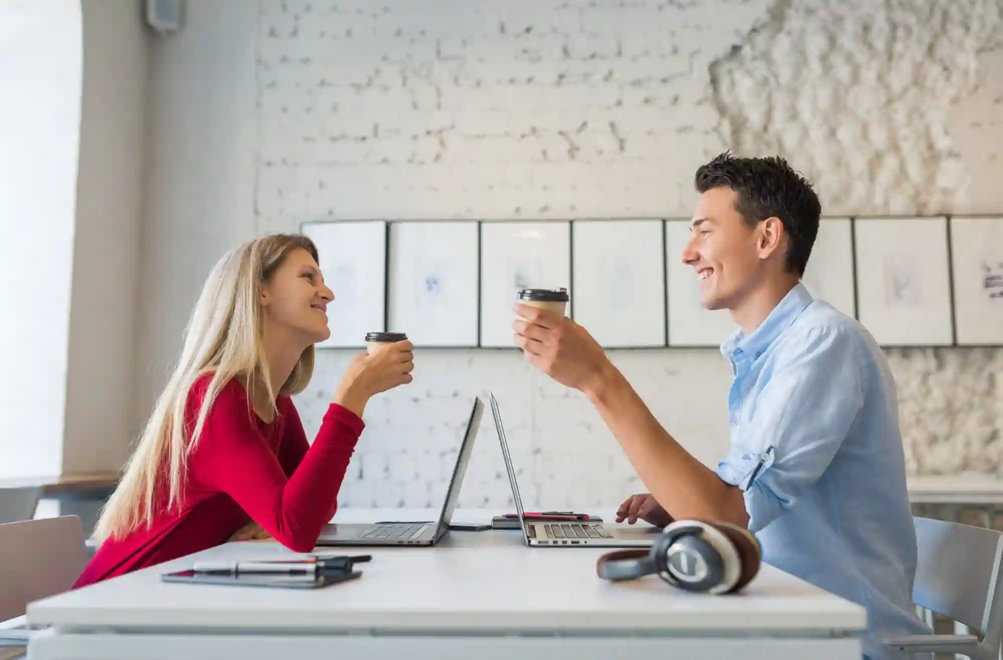 Man and woman smiling and raising coffee cups in a work or cafe setting.