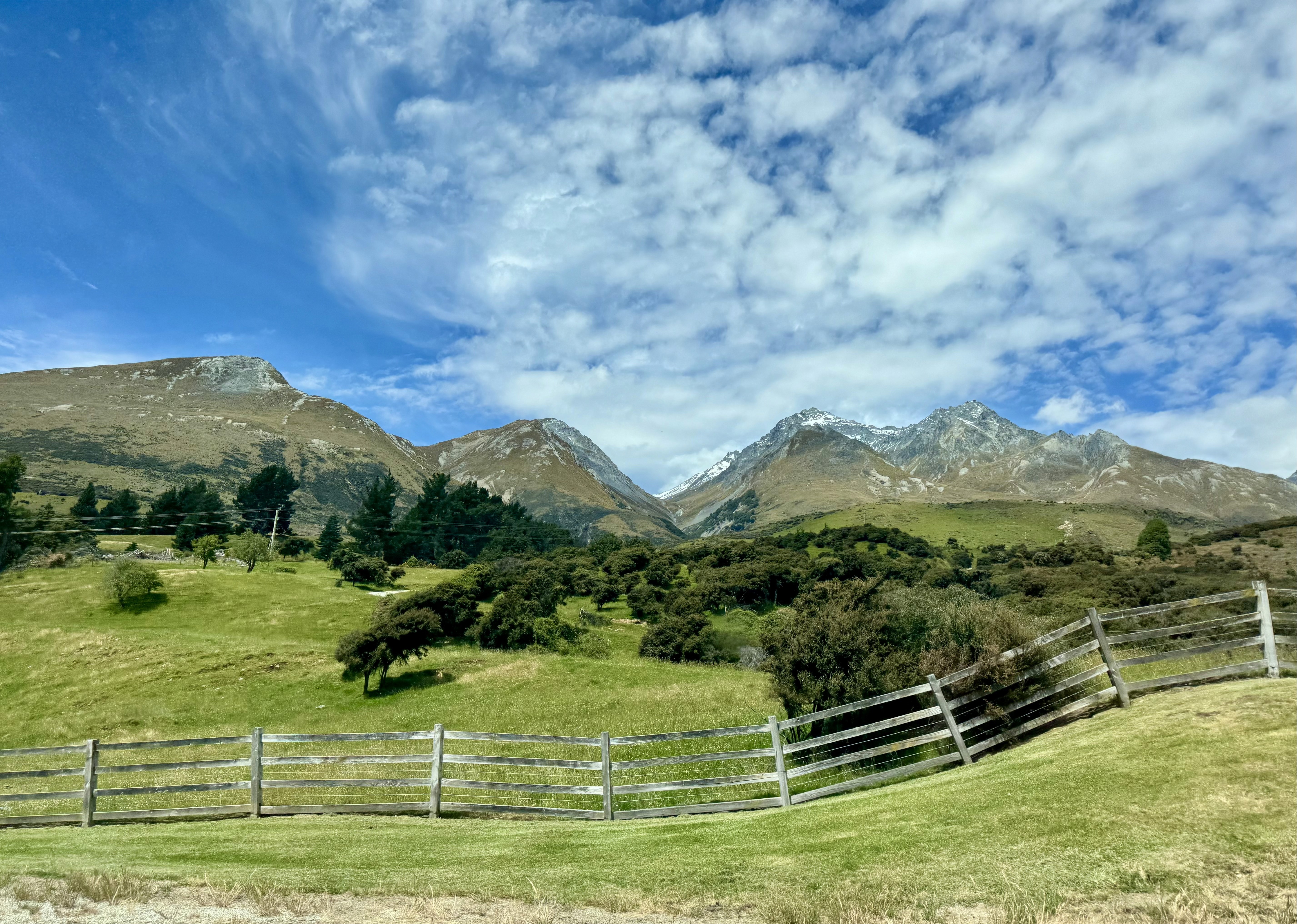 Rolling hills, lush green grass and snow capped mountains fenced across the road in Queenstown