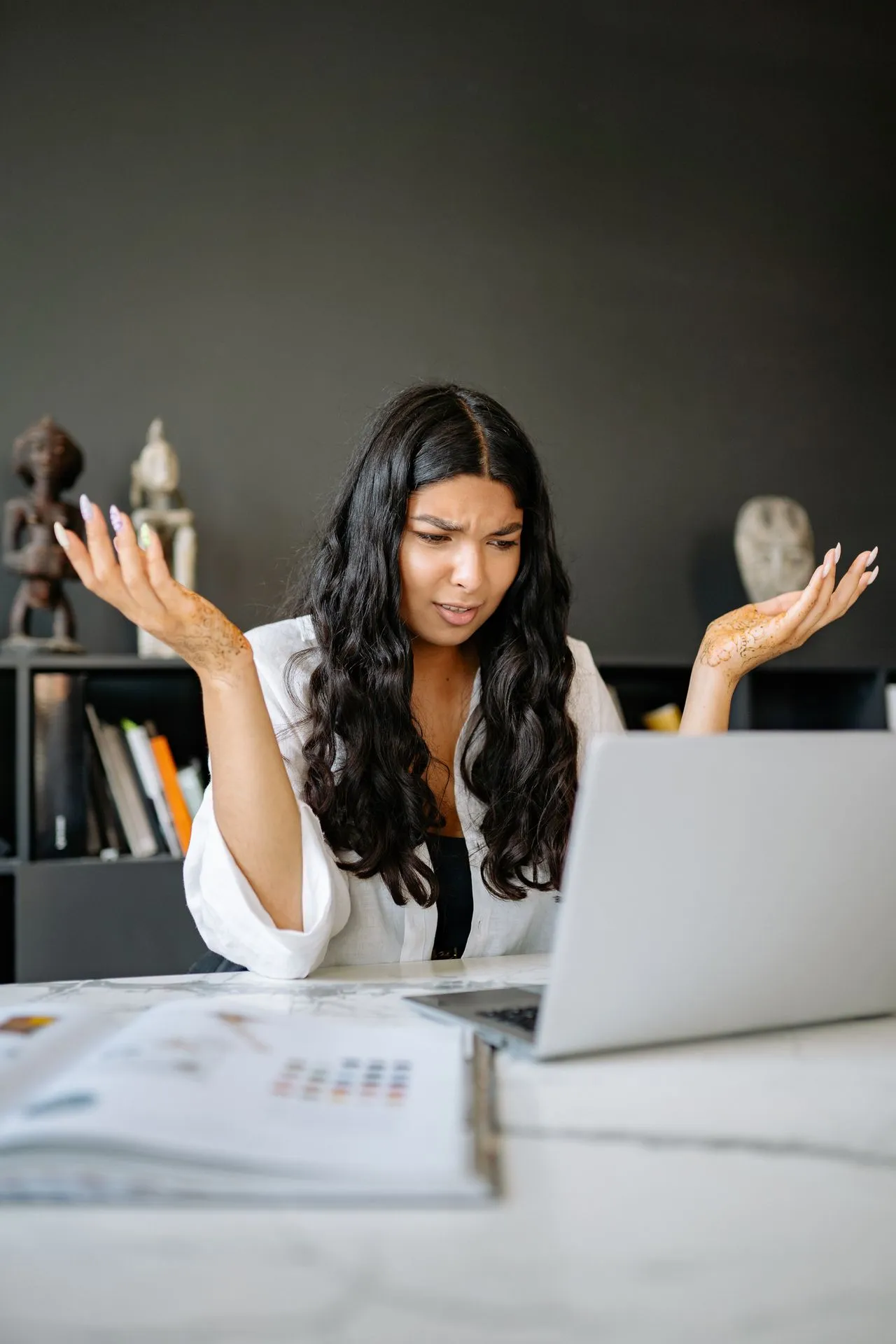 A woman with long, dark hair, wearing a white shirt, sits at a desk with an open laptop, looking confused as she gestures with her hands in frustration, surrounded by office decorations and papers.