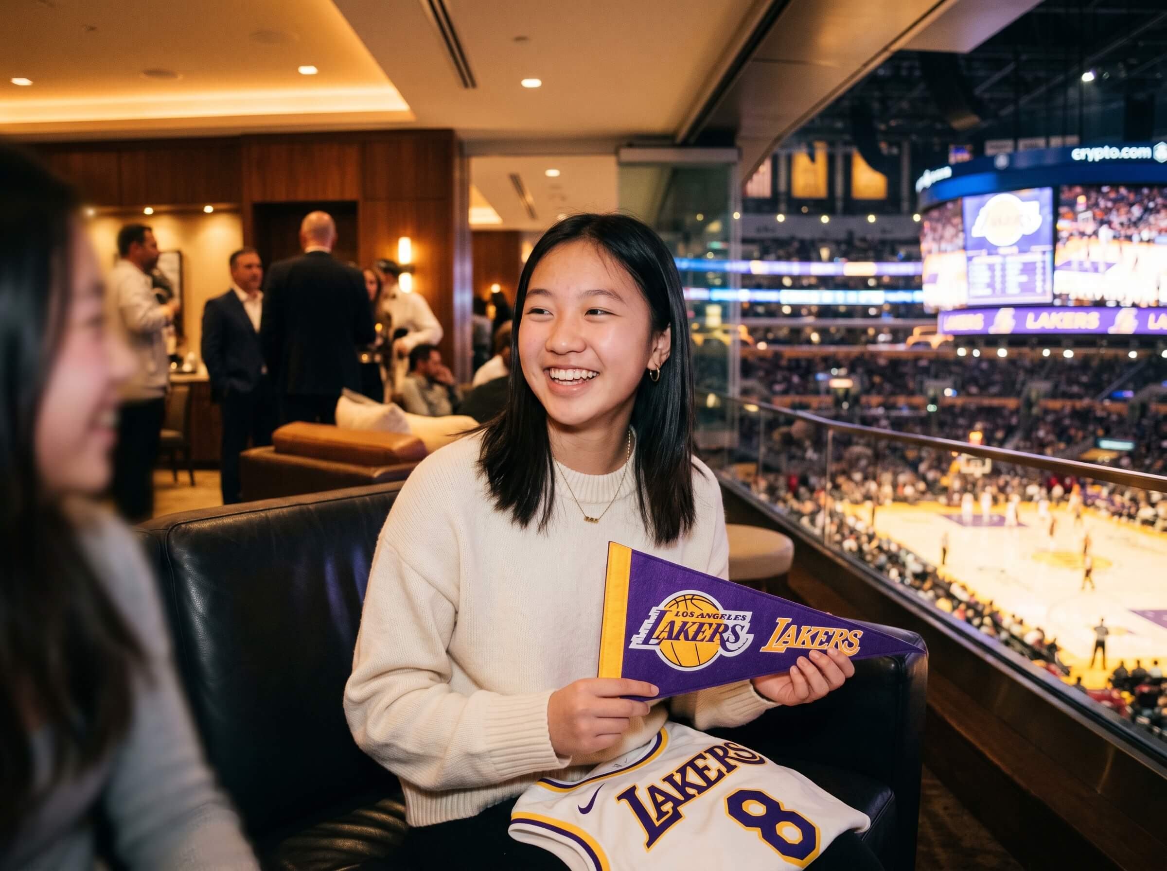 A person sits inside a luxury suite at a Lakers game, holding a Lakers pennant and jersey, with a view of the basketball court and large screen displaying the match in the background, under warm lighting with people interacting around.