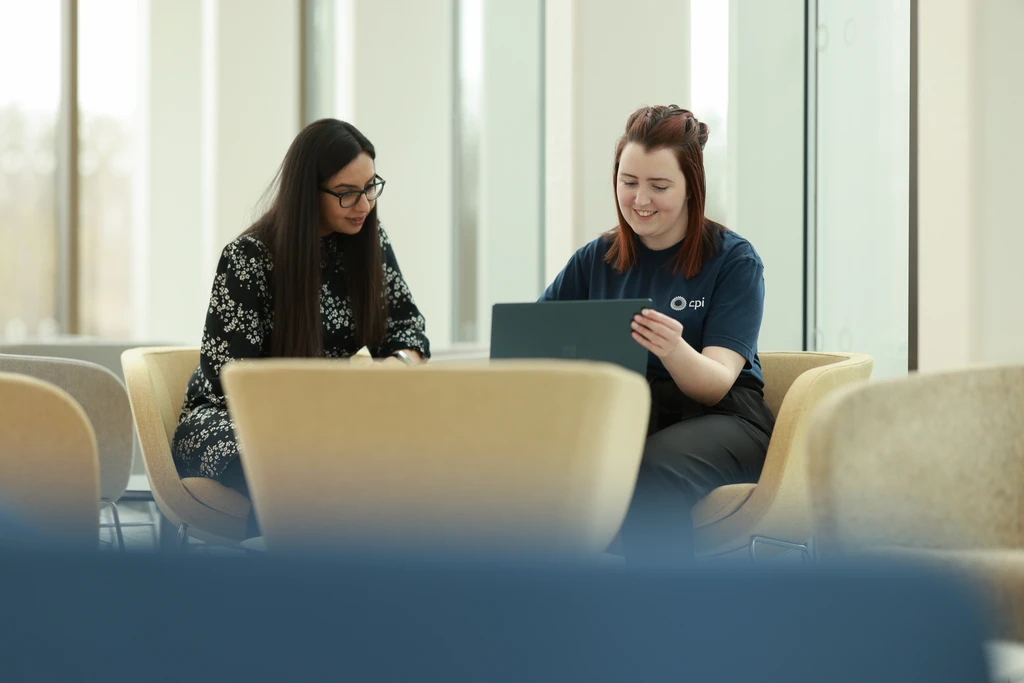 Two women sitting in a bright office lounge, smiling while looking at a tablet together.