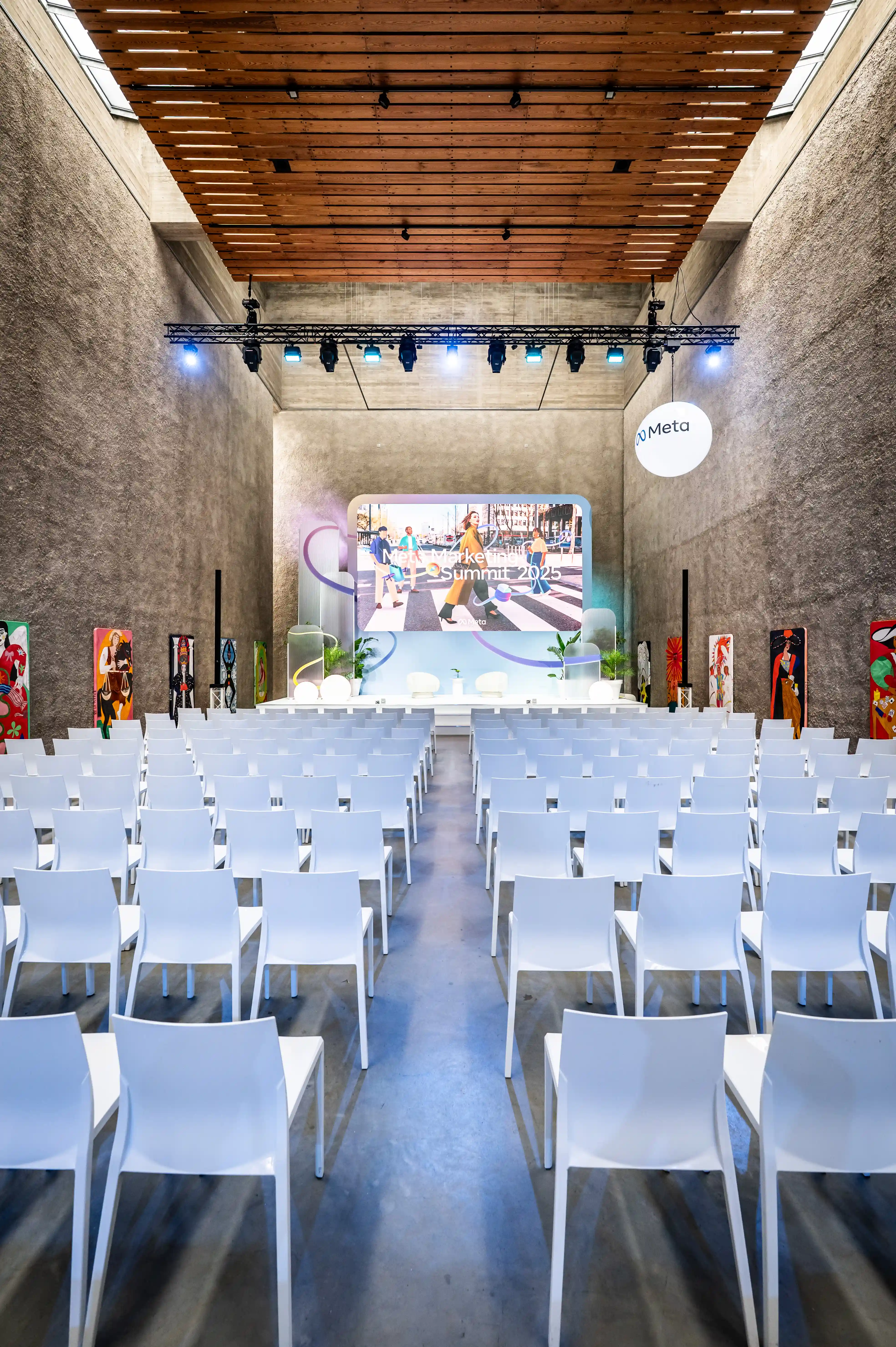 Tall concrete hall with white chairs facing the Meta Summit stage screen with colourful frame.