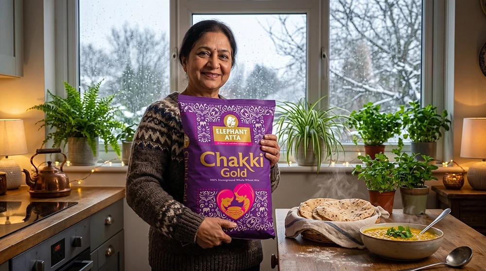South Asian woman smiling in cosy UK kitchen holding Elephant Atta Chakki Gold bag beside fresh rotis and dal