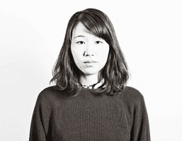 A black and white portrait of a young woman with shoulder-length dark hair and bangs. She wears a dark sweater with a simple crew neck and a thin, dark choker-style collar. Her expression is neutral, and her gaze is directed straight at the camera. The background is plain white with subtle texture.