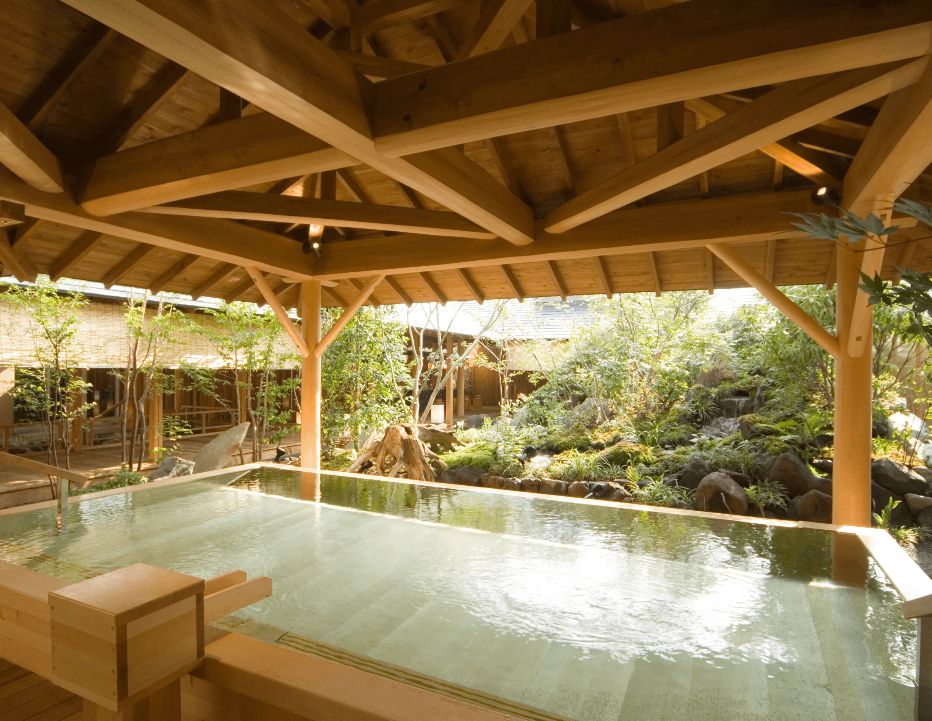 A large, rectangular Japanese onsen bath, made of light-colored wood, sits under a pavilion with an impressive exposed-beam timber roof. The open-sided structure offers a panoramic view of a tranquil Japanese courtyard garden with mossy rocks, trees, and covered walkways.