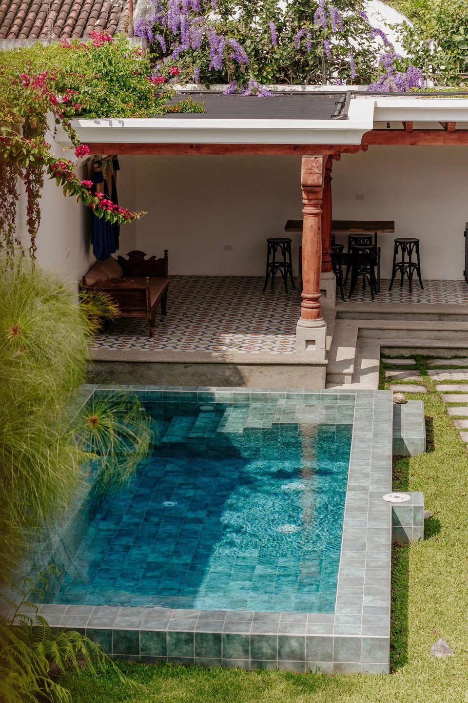 A serene backyard oasis featuring a small, tiled swimming pool with clear blue water, bordered by lush green grass and vibrant flowering plants, next to a covered patio with wooden columns and bar stools, surrounded by rustic architectural elements.