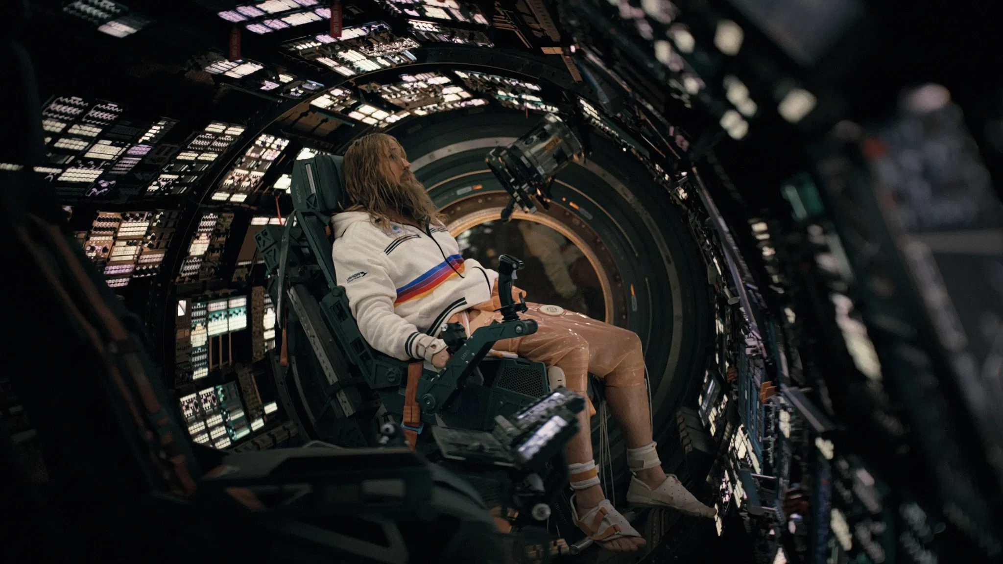 A person with long hair and a beard is seated in a high-tech spacecraft cockpit, gazing at multiple illuminated control panels, conveying a futuristic space exploration theme.