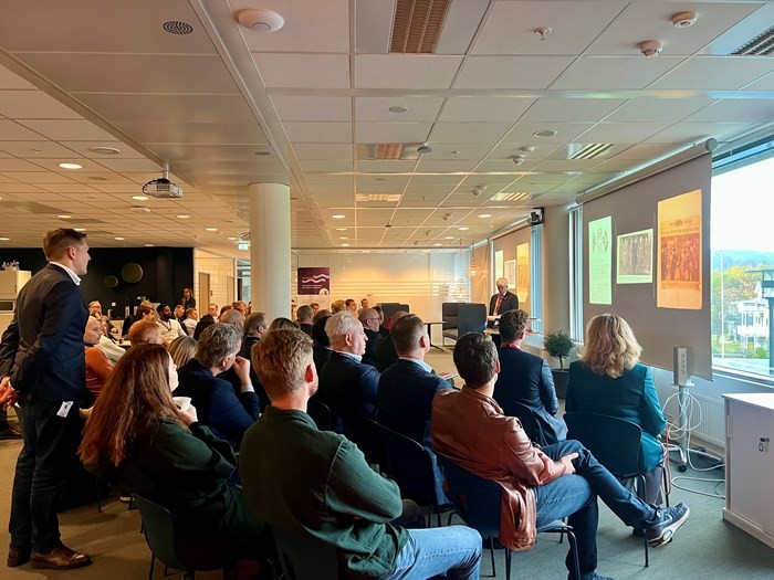 A group of people seated in a modern meeting room, attentively facing a speaker near a window.