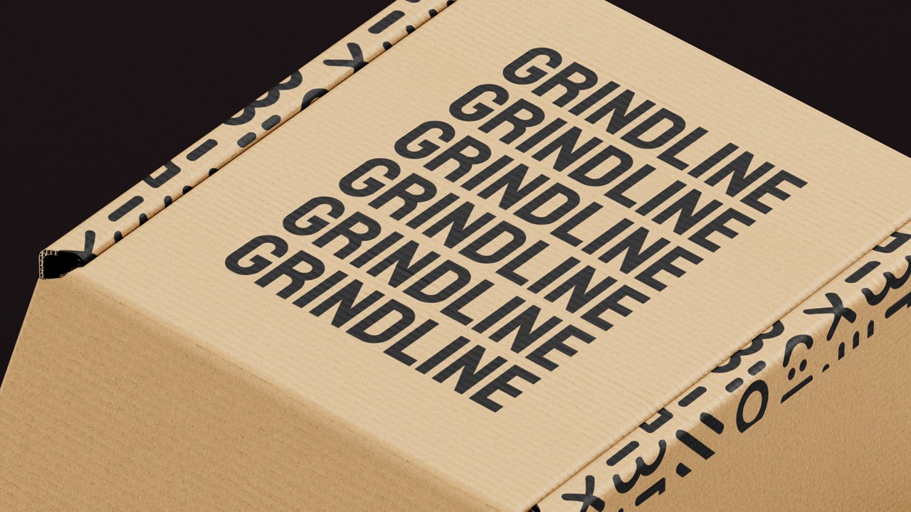 Corrugated cardboard box on black showing multiple stacked “GRINDLINE” wordmarks and black icon-patterned tape along the edges.