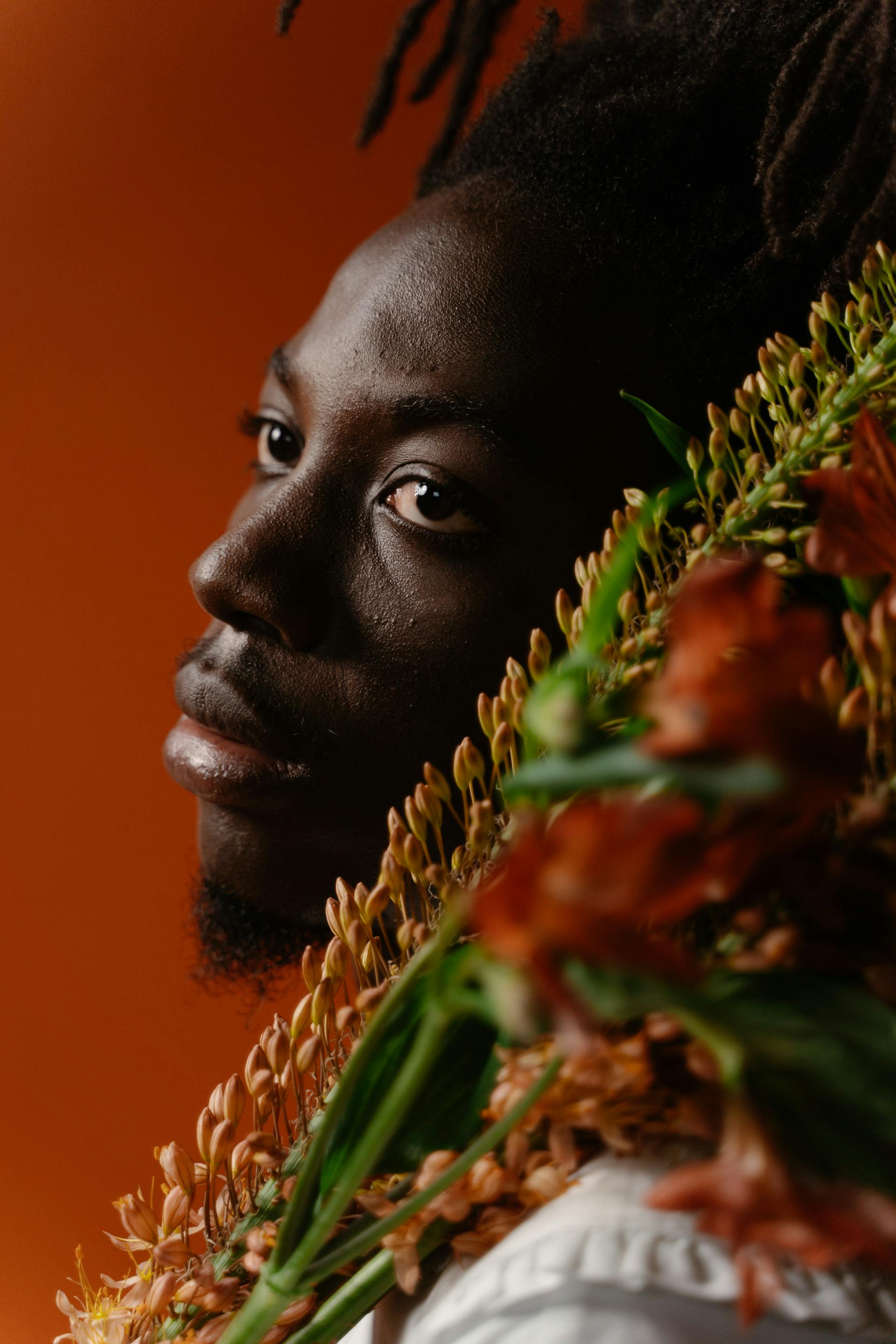 A profile portrait of a man looking over his shoulder, partially framed by a bunch of reddish-orange flowers against a solid orange backdrop.