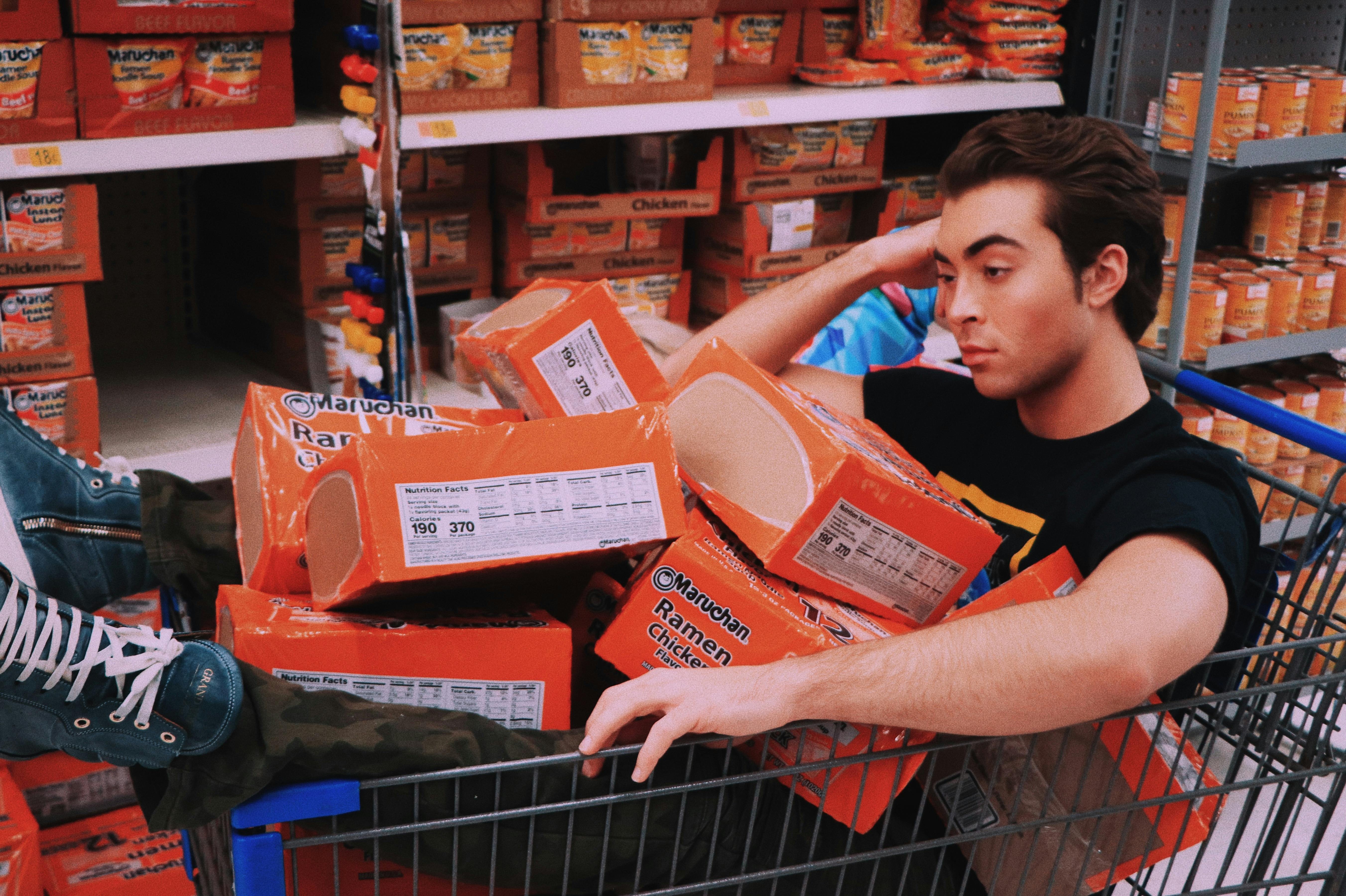 A young man sitting in a shopping trolley filled with Maruchan Ramen boxes in a supermarket aisle, surrounded by shelves stocked with packaged goods – illustrating the influence of brand experience on customer behaviour in retail environments.