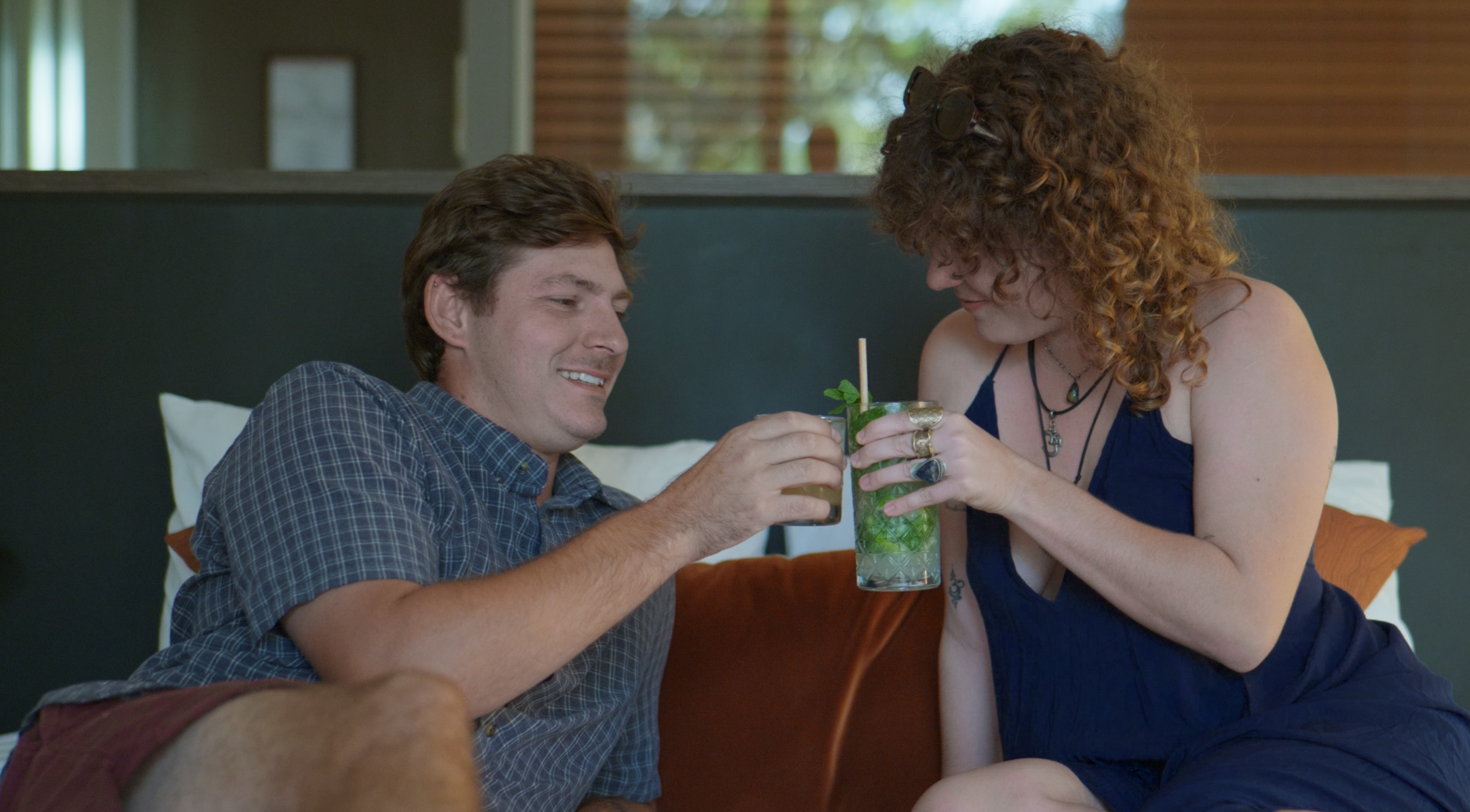 A couple relaxes on a cozy couch, smiling as they clink glasses filled with refreshing cocktails, framed by a stylish indoor setting.