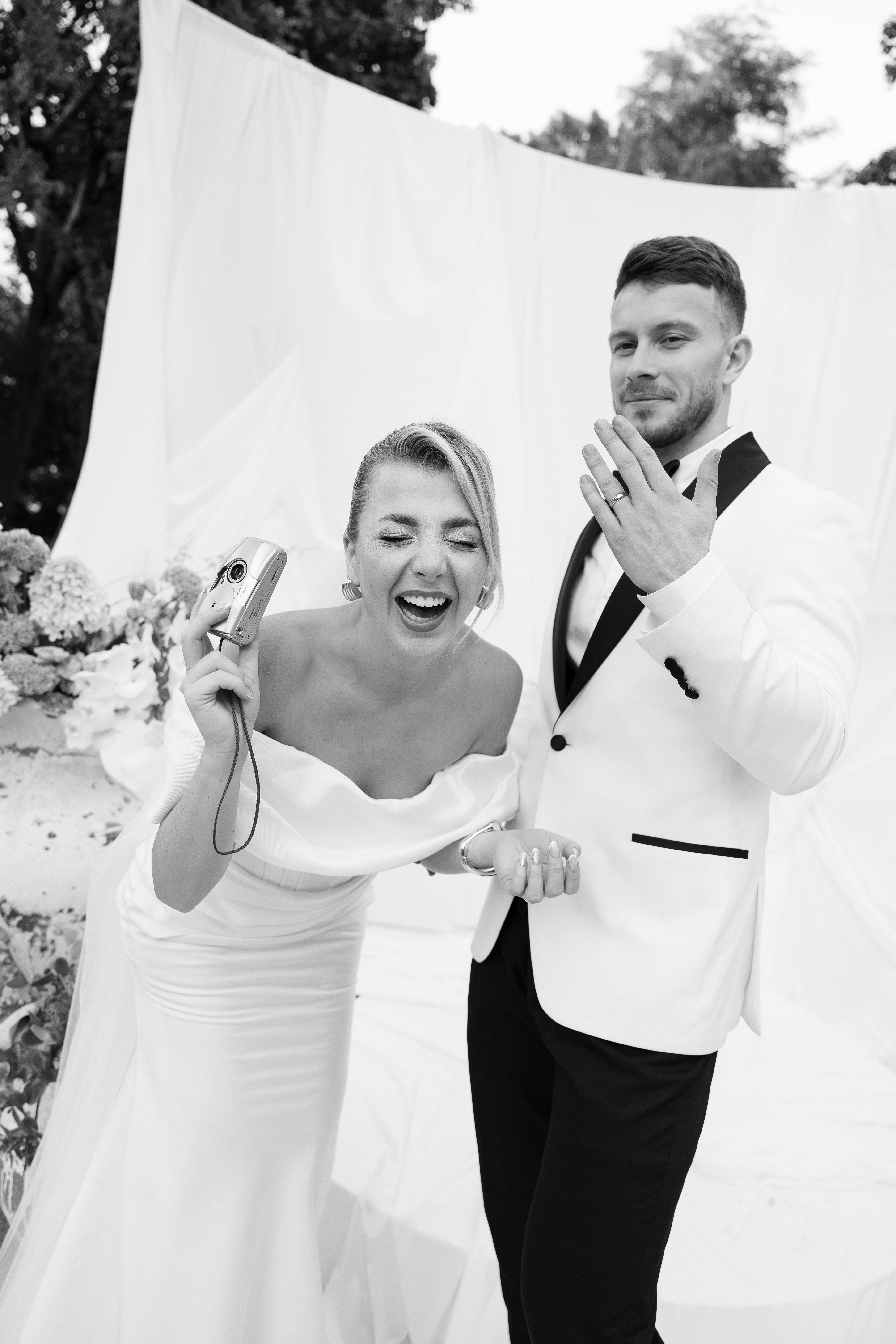 Bride and groom laughing and holding drinks — candid joy in a summer setting.