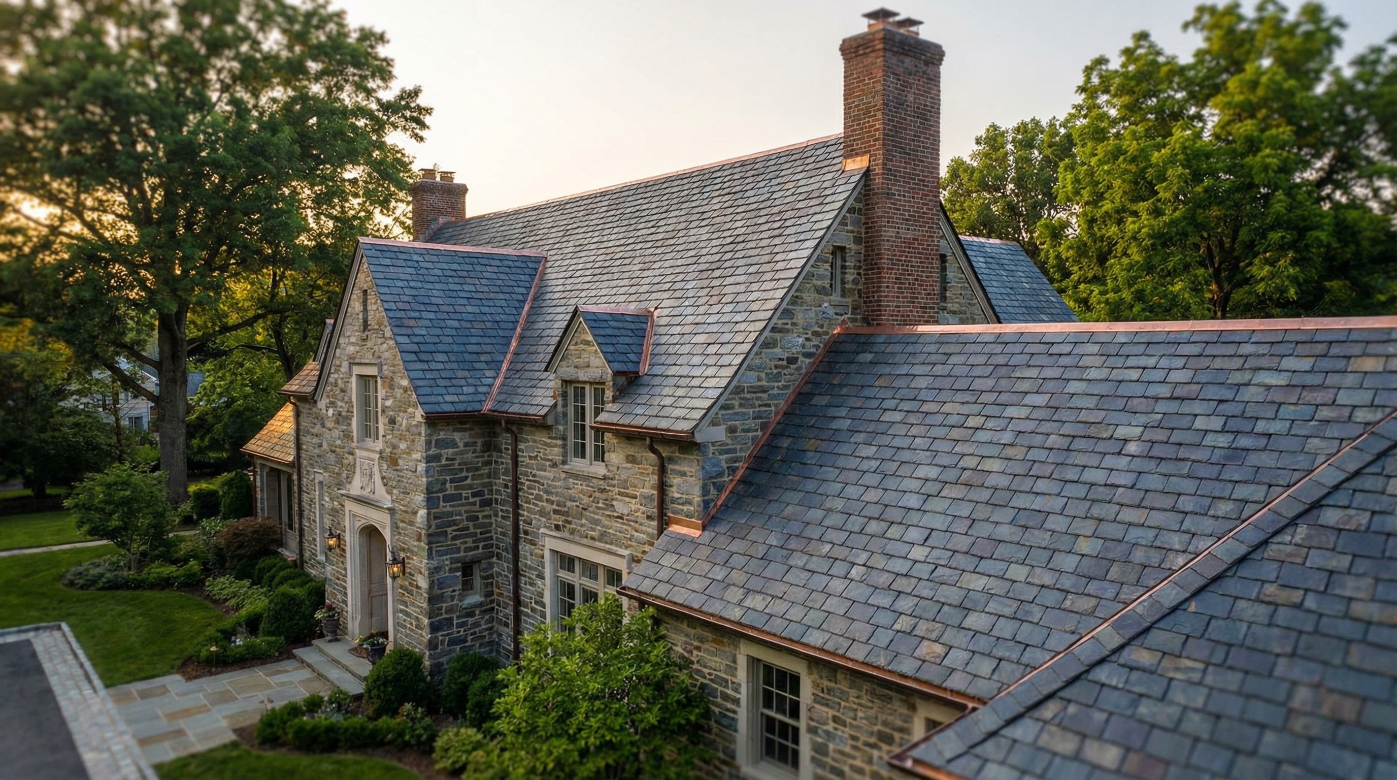 Stone house with a slate roof, brick chimney, and lush greenery in a residential area. Architecture and real estate.