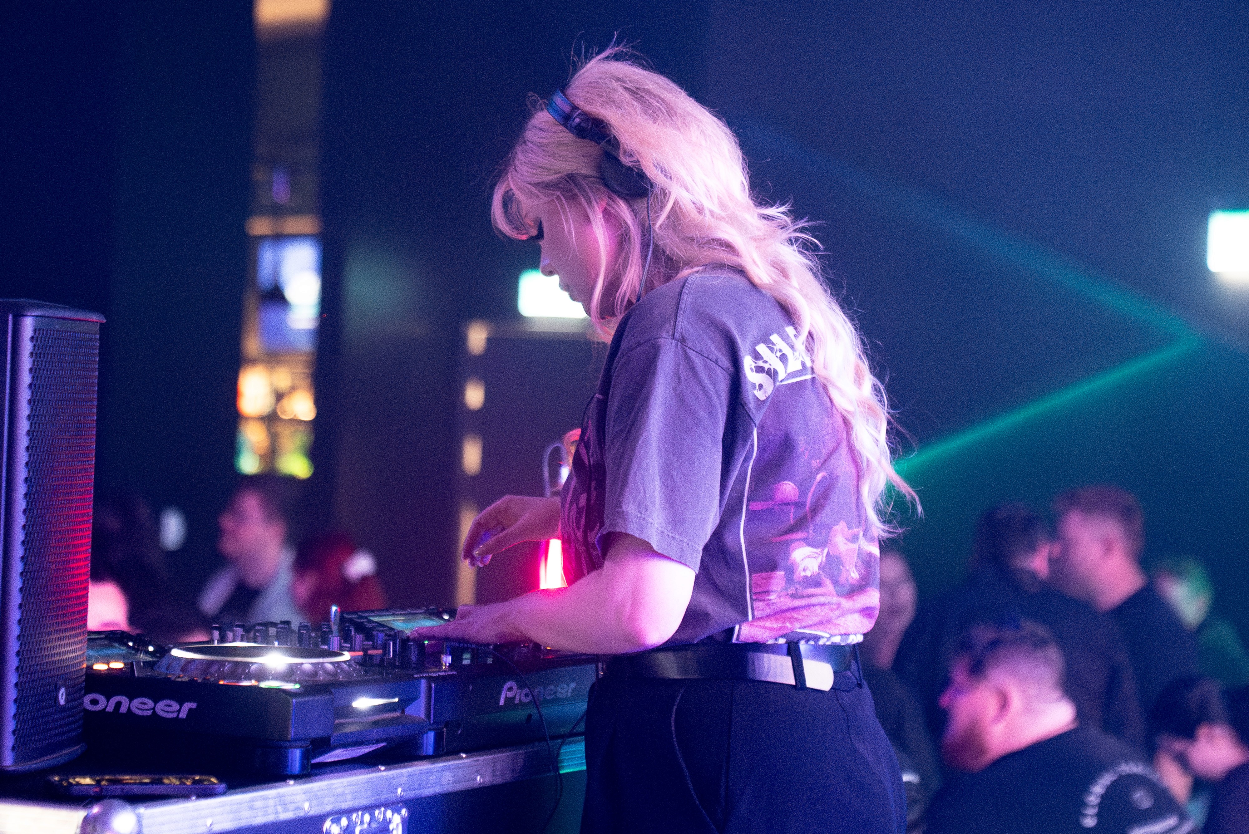 woman standing behind dj deck looking down at the mixer while people dance in background