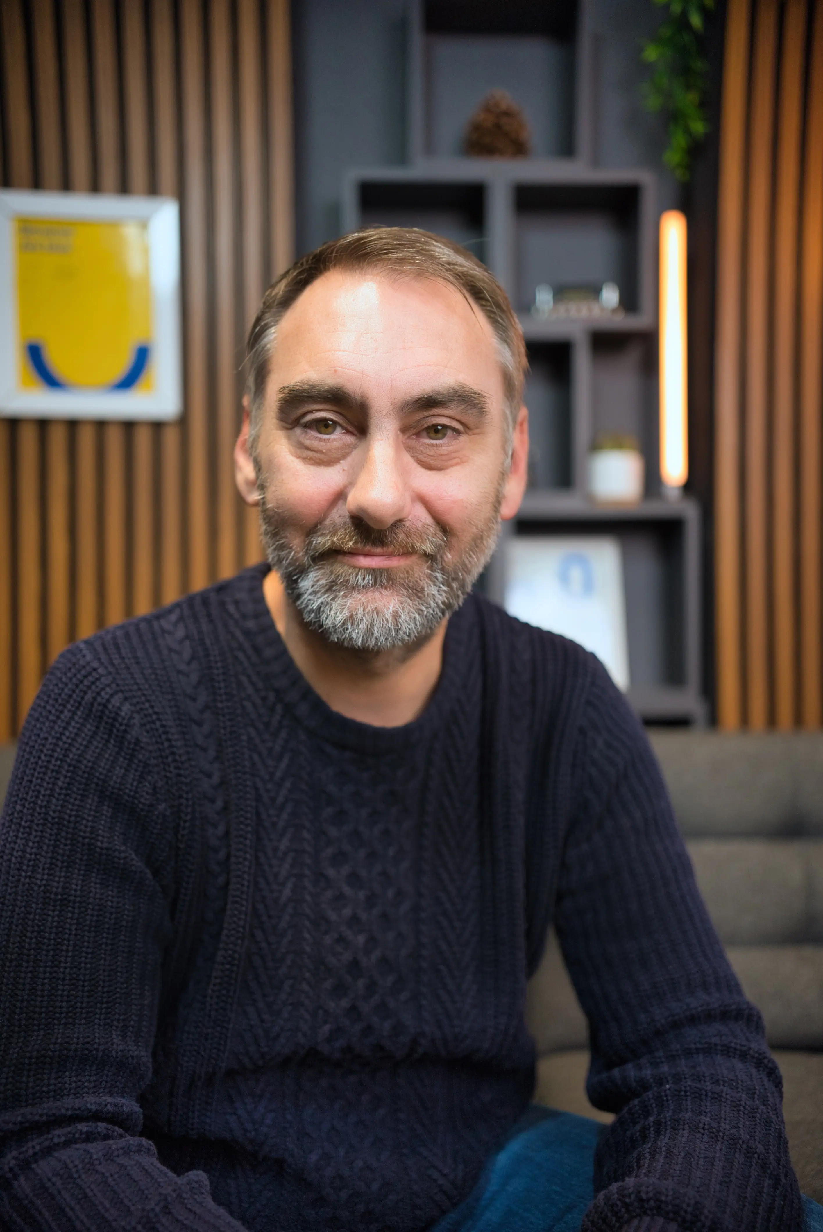A man with a beard and a dark sweater sits on a grey sofa against a wooden panel background, with various framed pictures on the wall.