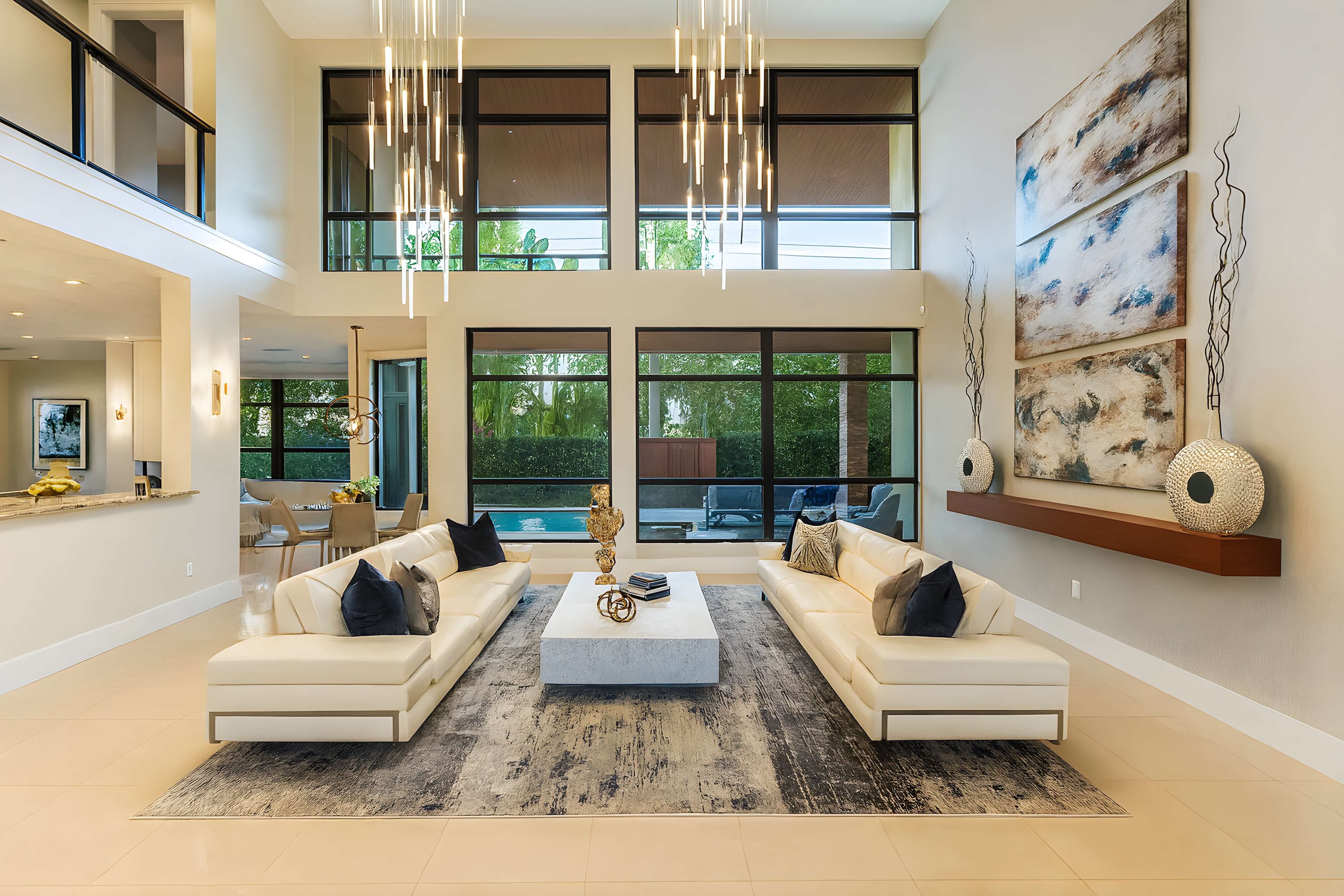 Spacious modern living room with double-height ceilings, two white leather sofas facing a marble coffee table, large abstract wall art, statement pendant lighting, and expansive windows overlooking a pool.