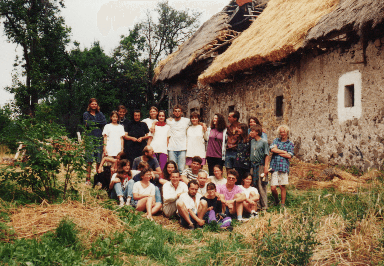 A young person with curly hair smiles at the camera while sitting in front of a laptop against a dark background.