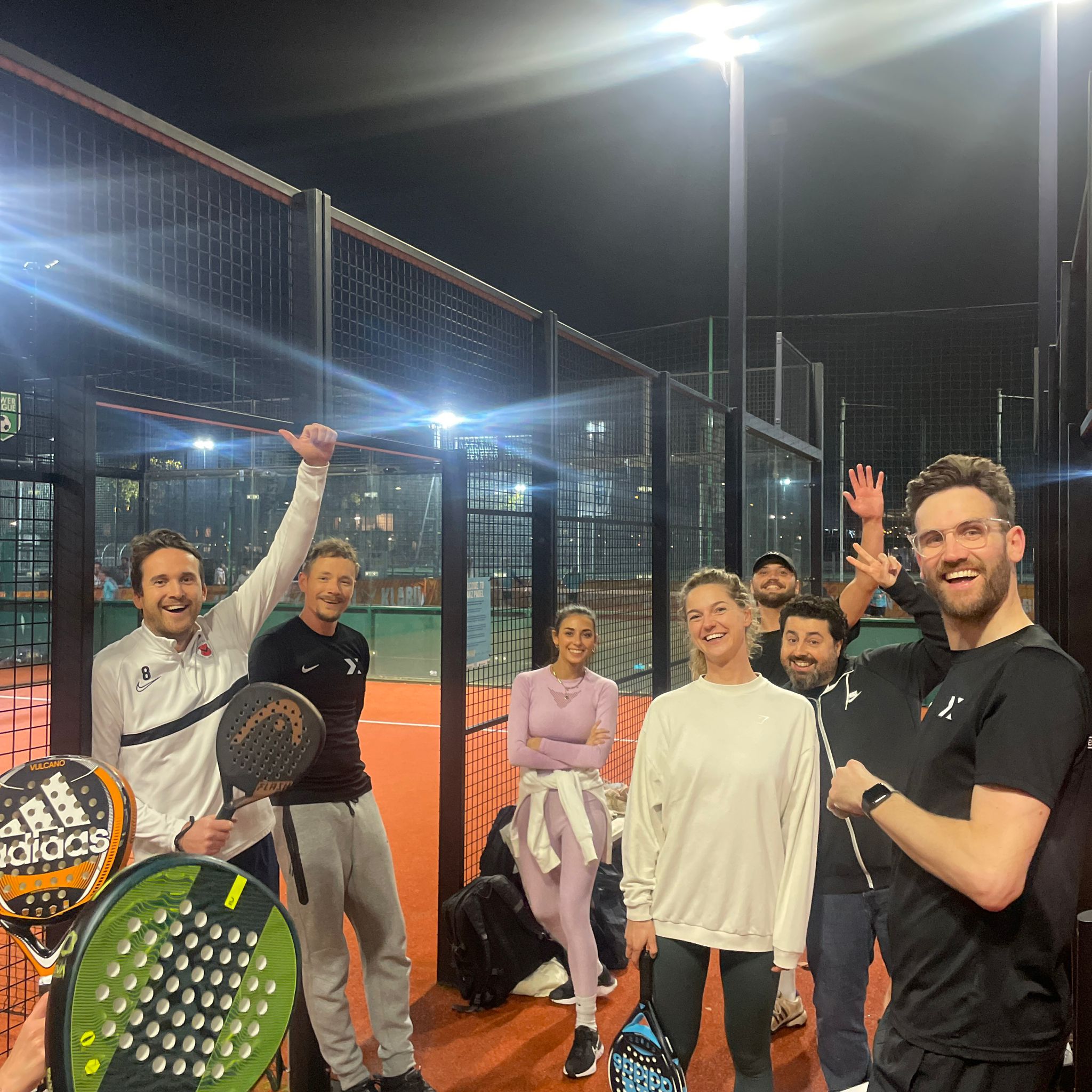A group of seven people poses on a brightly lit tennis court, celebrating after a game.
