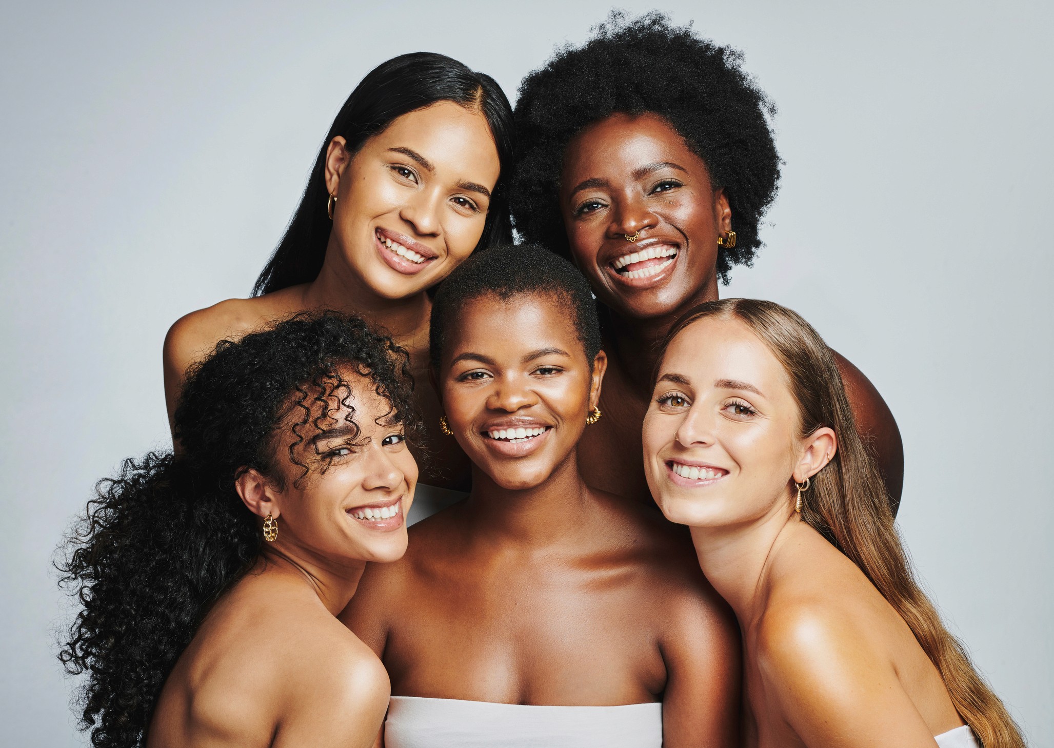 A group of five diverse women is posing closely together, smiling warmly against a neutral background. They have radiant, natural skin and are wearing minimal makeup, emphasizing beauty in all shades and textures. Their expressions exude confidence, happiness, and a strong sense of unity, celebrating inclusivity and diversity in beauty.
