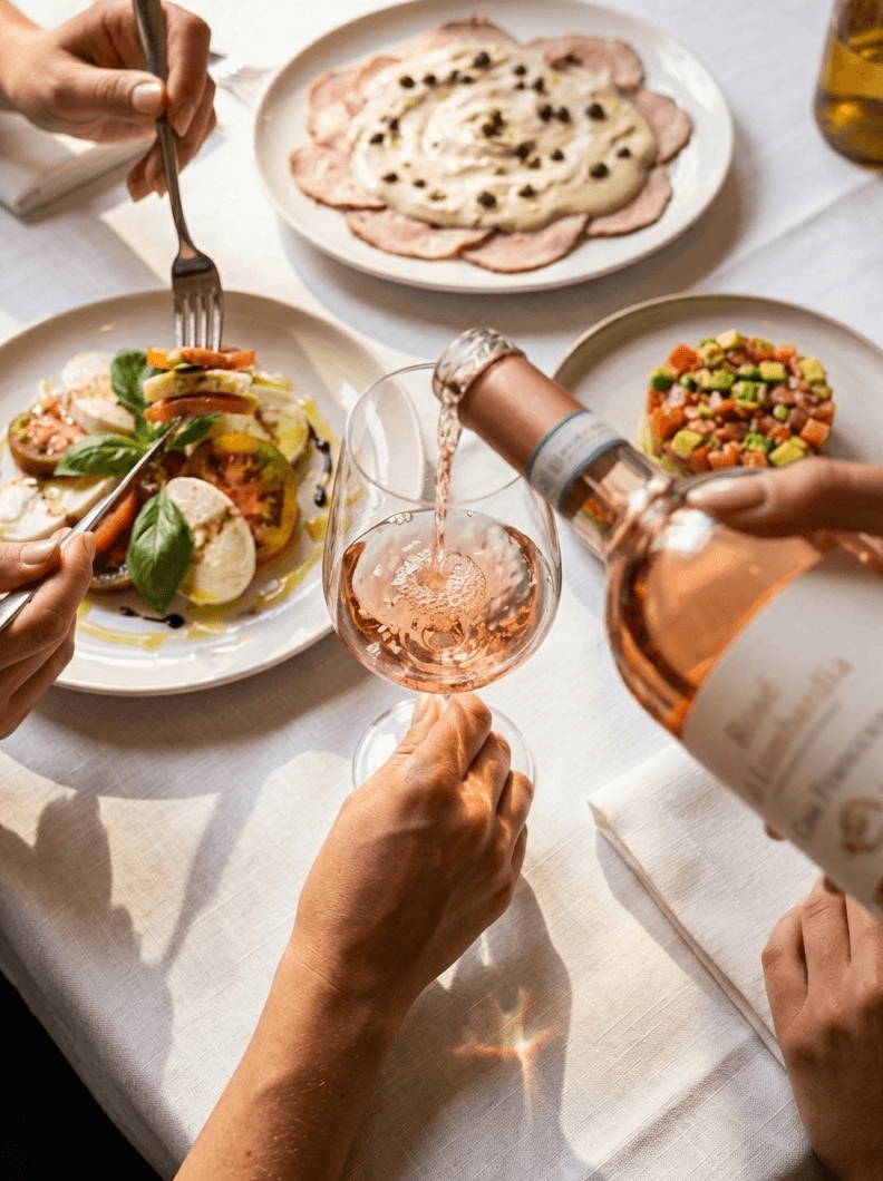 Vino rosado sirviéndose en una mesa con platos italianos, entre ellos una ensalada caprese y pequeños platos para compartir.