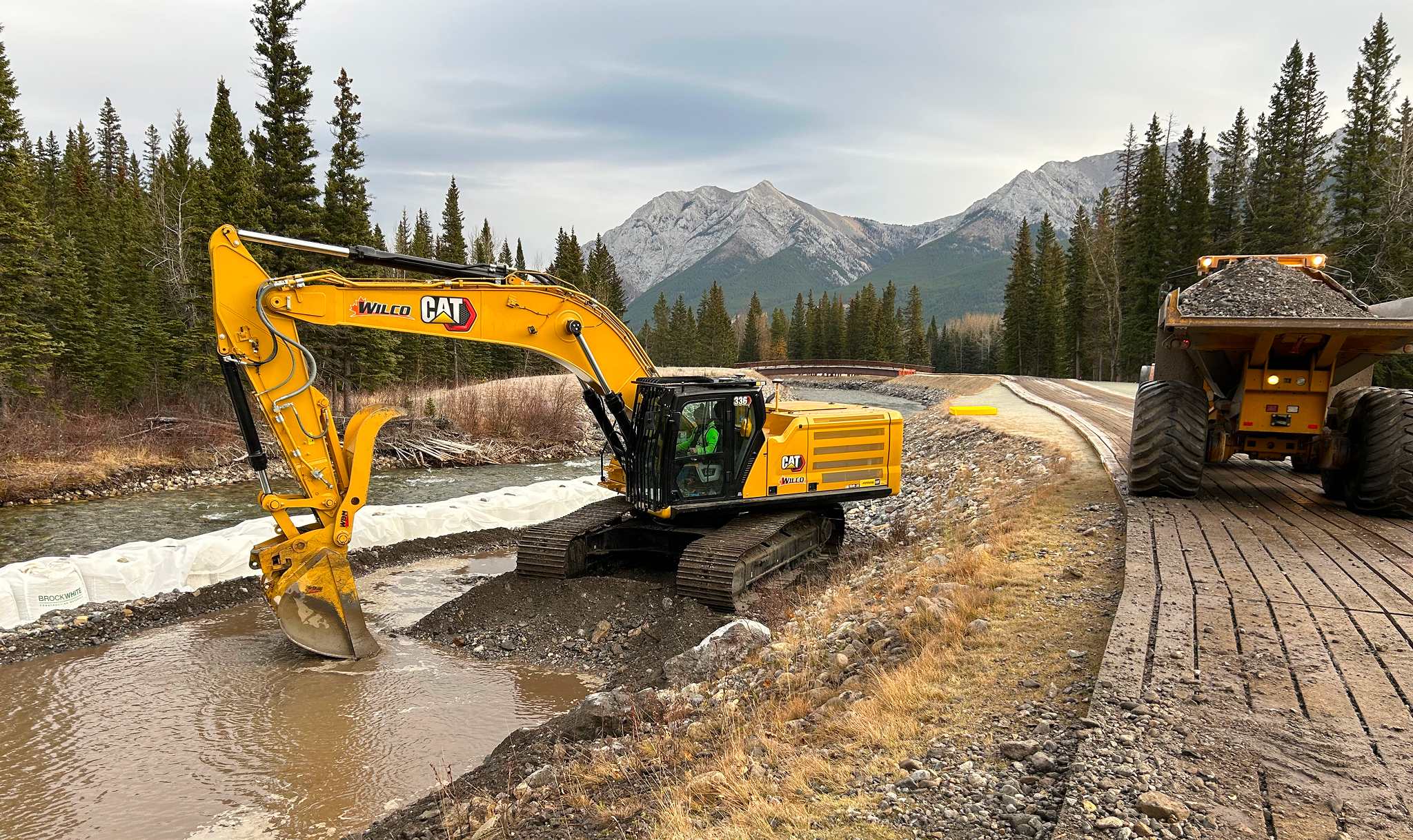 Excavator removing sediment near Kananaskis River with temporary timber mat access road and mountain backdrop