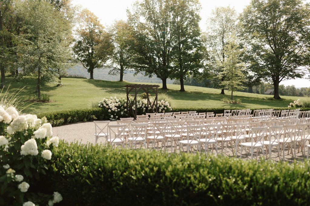 Wide view of an outdoor wedding ceremony site with rows of white chairs, a wooden pergola, hydrangea hedges, and rolling green hills