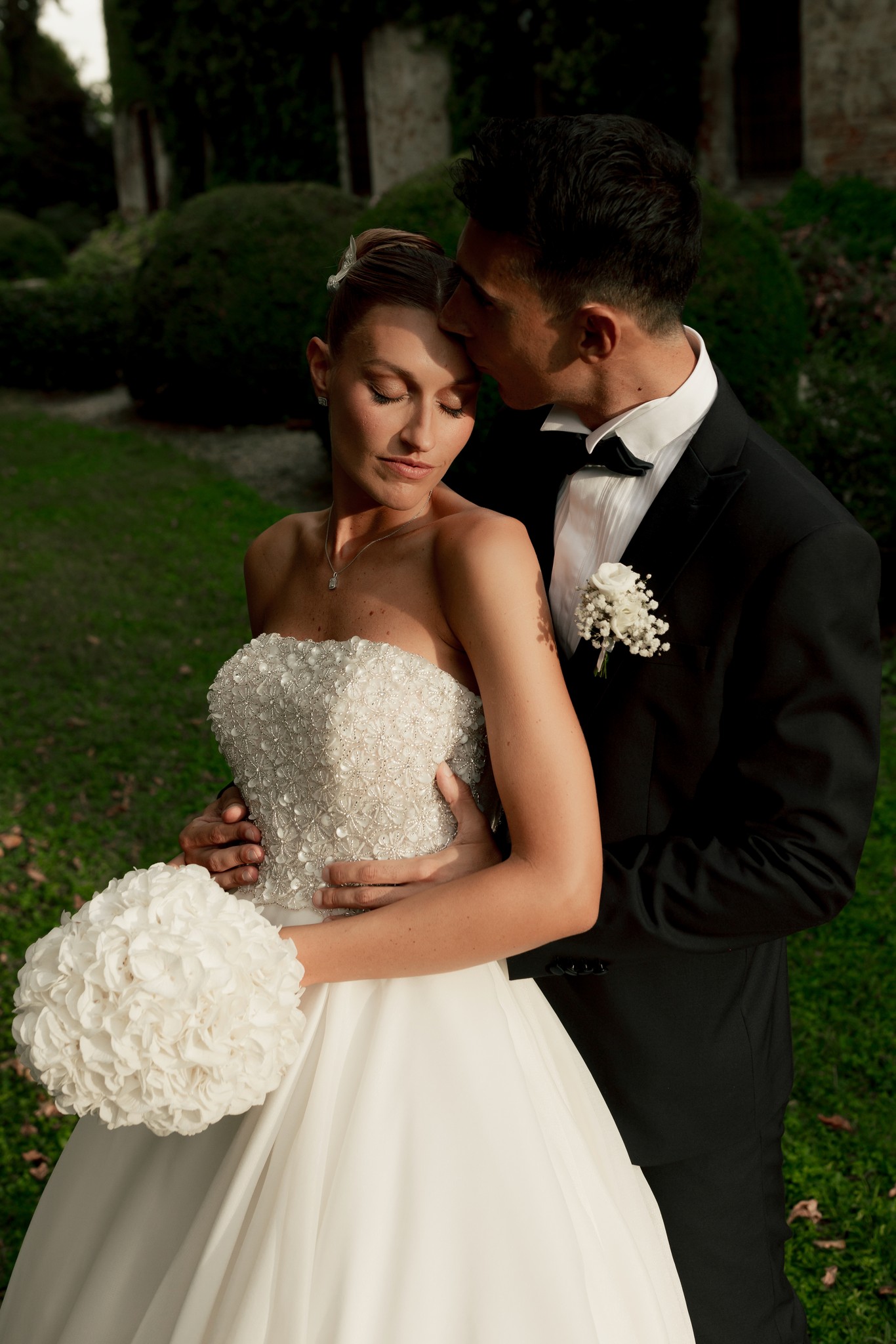 Eleonora e Fabio si abbracciano nel parco di Villa Ferrari, lei con bouquet bianco, lui in smoking con spilla floreale