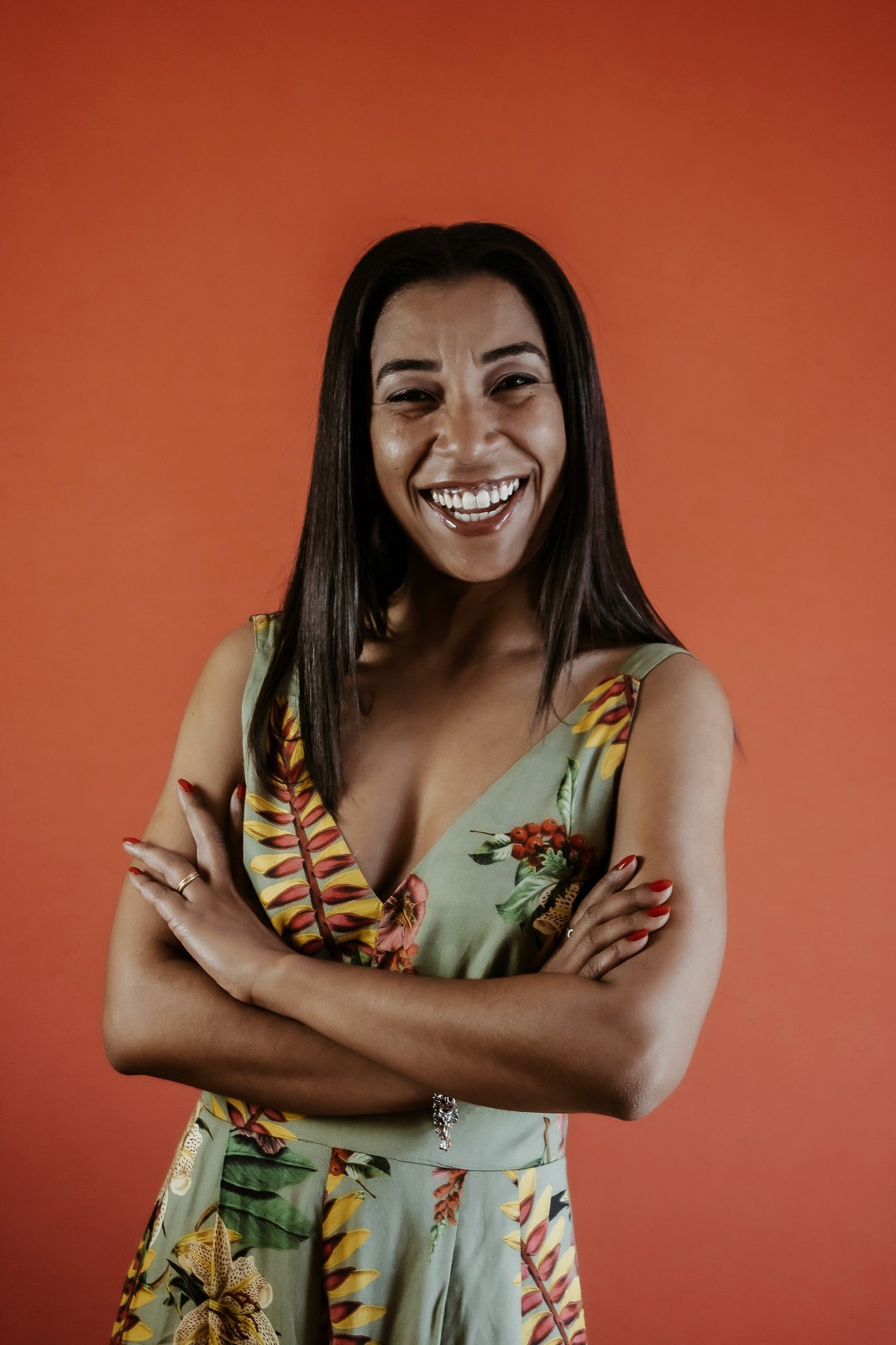 Mulher negra de cabelos compridos, sorrindo para a tela, de vestido estampado verde no fundo alaranjado