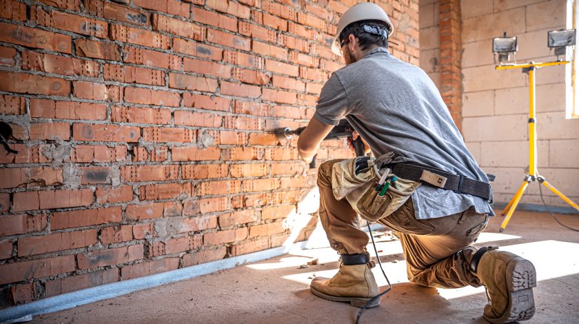 A construction worker in a gray helmet operates a power drill while kneeling on a concrete floor, focused on a brick wall, with a tool belt and work boots, under the illumination of standing work lights.