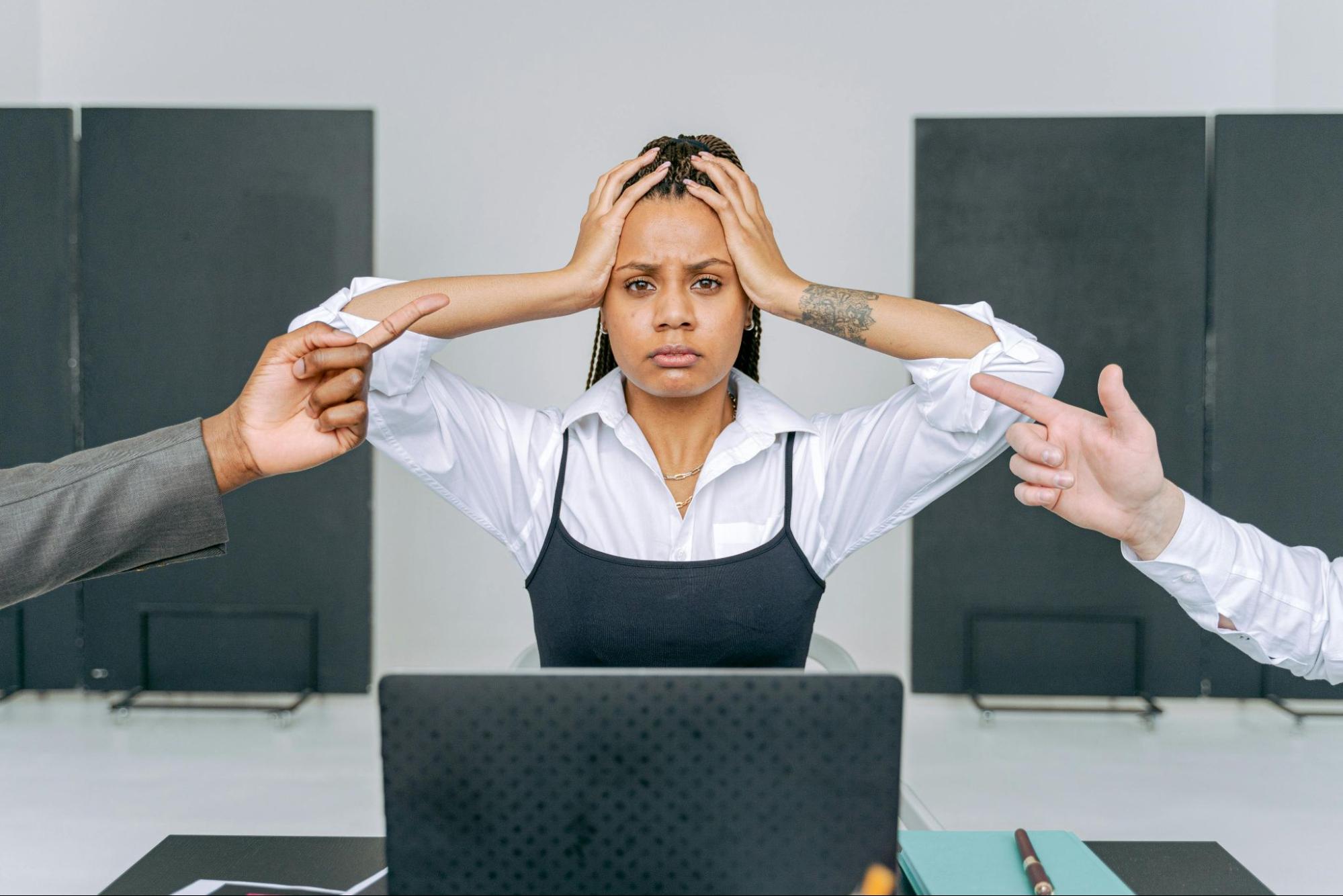 Lady in a white shirt holding her head with two hands. She looks upset or stressed. She has a laptop in front of her and there are two hands pointing from either side of the screen as if to be in an argument.