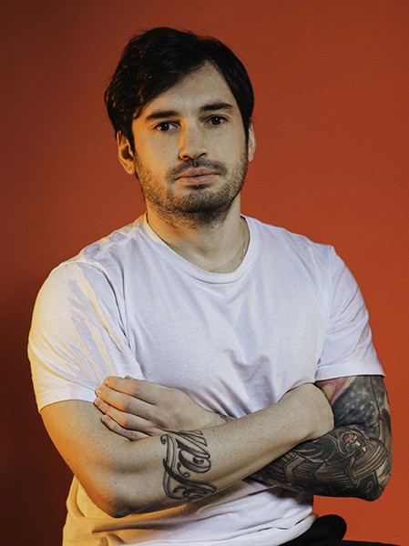 A man with dark hair and a beard stands against a solid orange-brown background. He wears a plain white t-shirt and has his arms crossed over his chest. He has a tattoo on his left forearm that includes cursive lettering and a design that includes what appears to be a stylized eagle or bird. His expression is neutral.