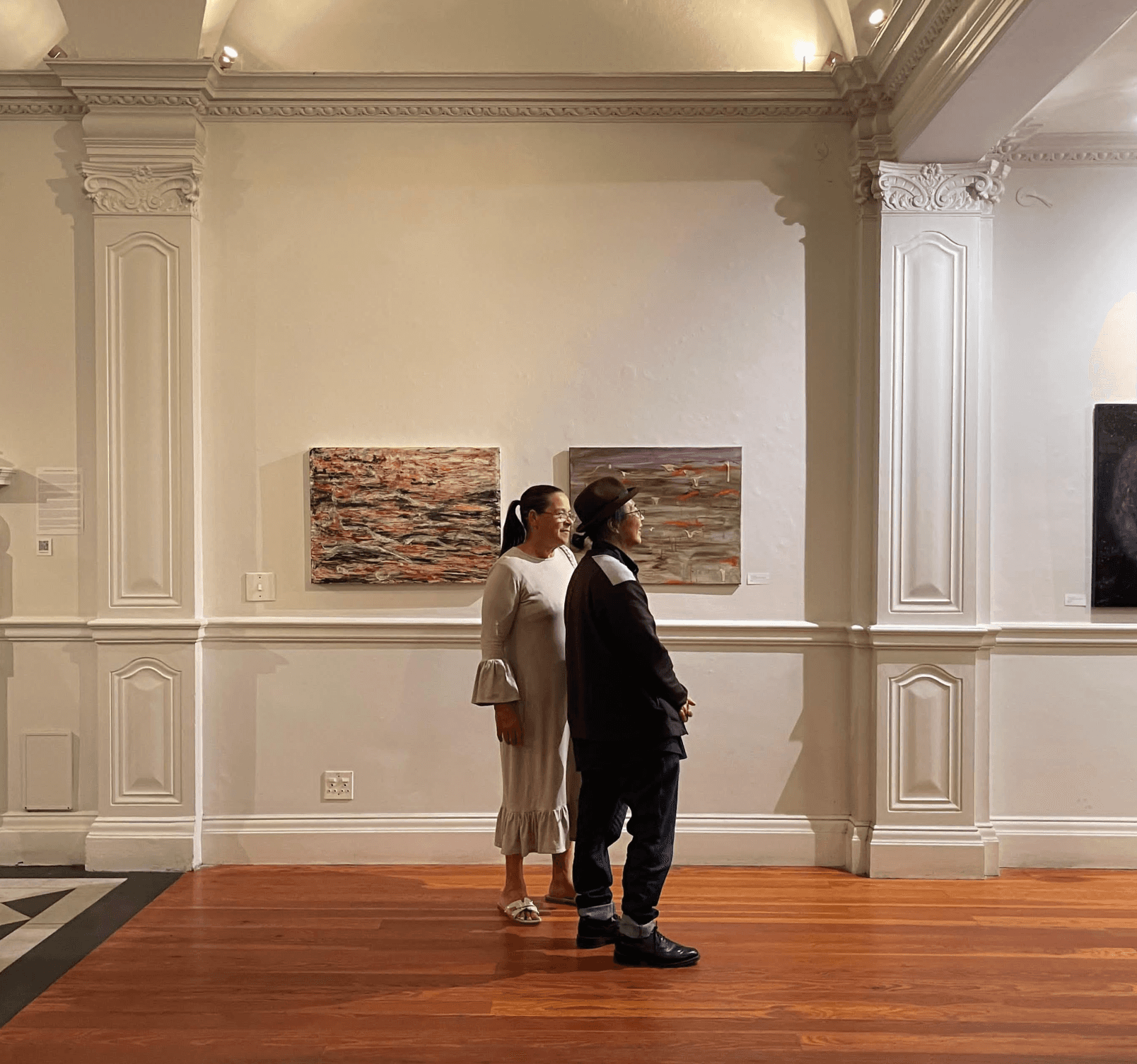 two people facing to the right, standing in front of two arworks