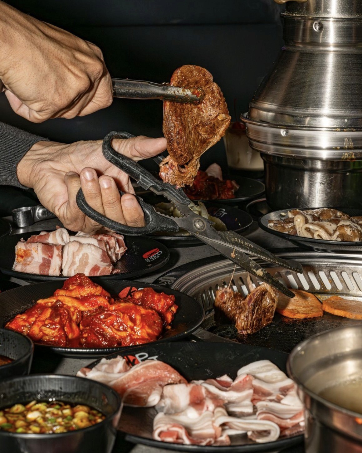 "An inviting table setup at KPOT Korean BBQ featuring sizzling Korean barbecue meats, steaming hot pot broth, fresh vegetables, and homemade sauces."