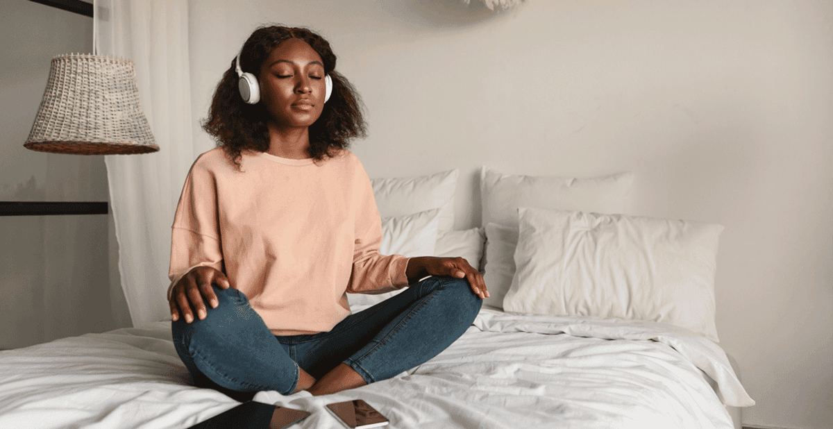 Woman sitting cross-legged on a bed, meditating with headphones on.