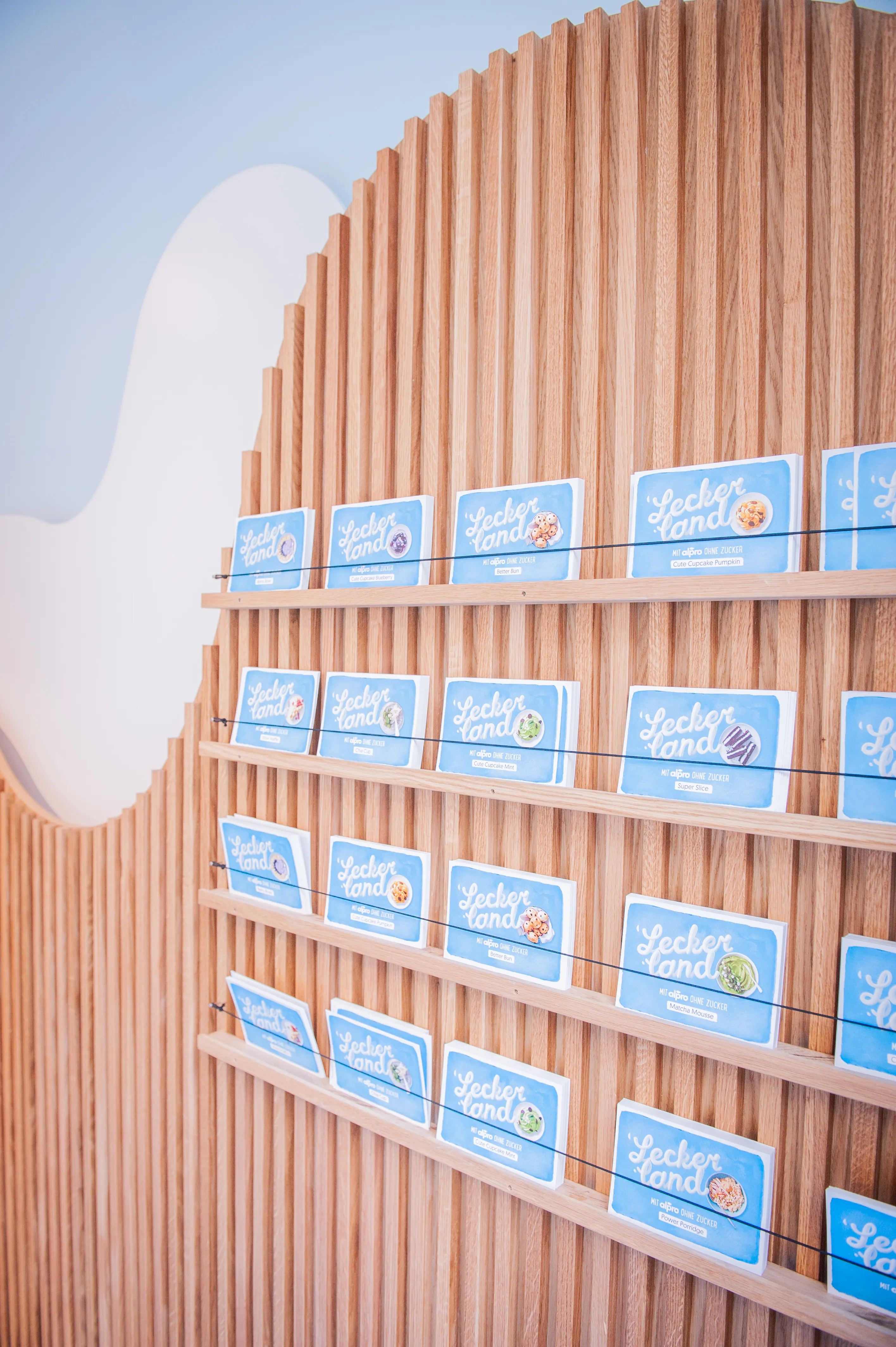 Curved wood-slat wall with rows of blue Leckerland recipe cards on narrow shelves at the Alpro pop-up.