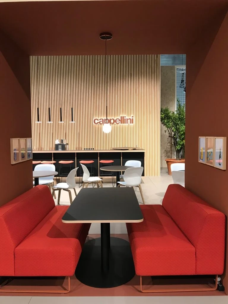 Seating nook at the HAWORTH ORGATEC stand with two red facing sofas and a black table in a rust-red frame, with the cappellini bar behind.