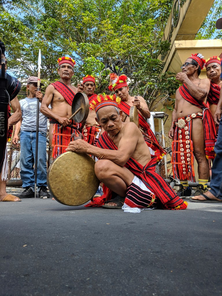 A group of indigenous men from the Philippines, dressed in traditional red and black woven attire, perform a cultural dance with gongs and spears during a vibrant outdoor festival.