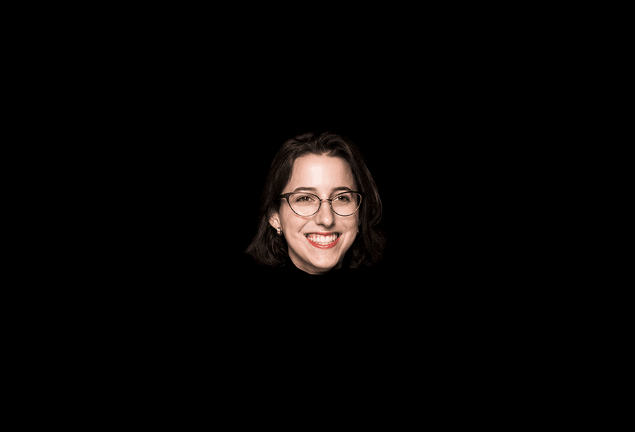 A woman with shoulder-length dark hair and round glasses is centered against a solid black background. She is smiling, showing her teeth, and her gaze is directed toward the camera. She wears a dark top and small earrings. The lighting is focused on her face, leaving the background dark and undefined.