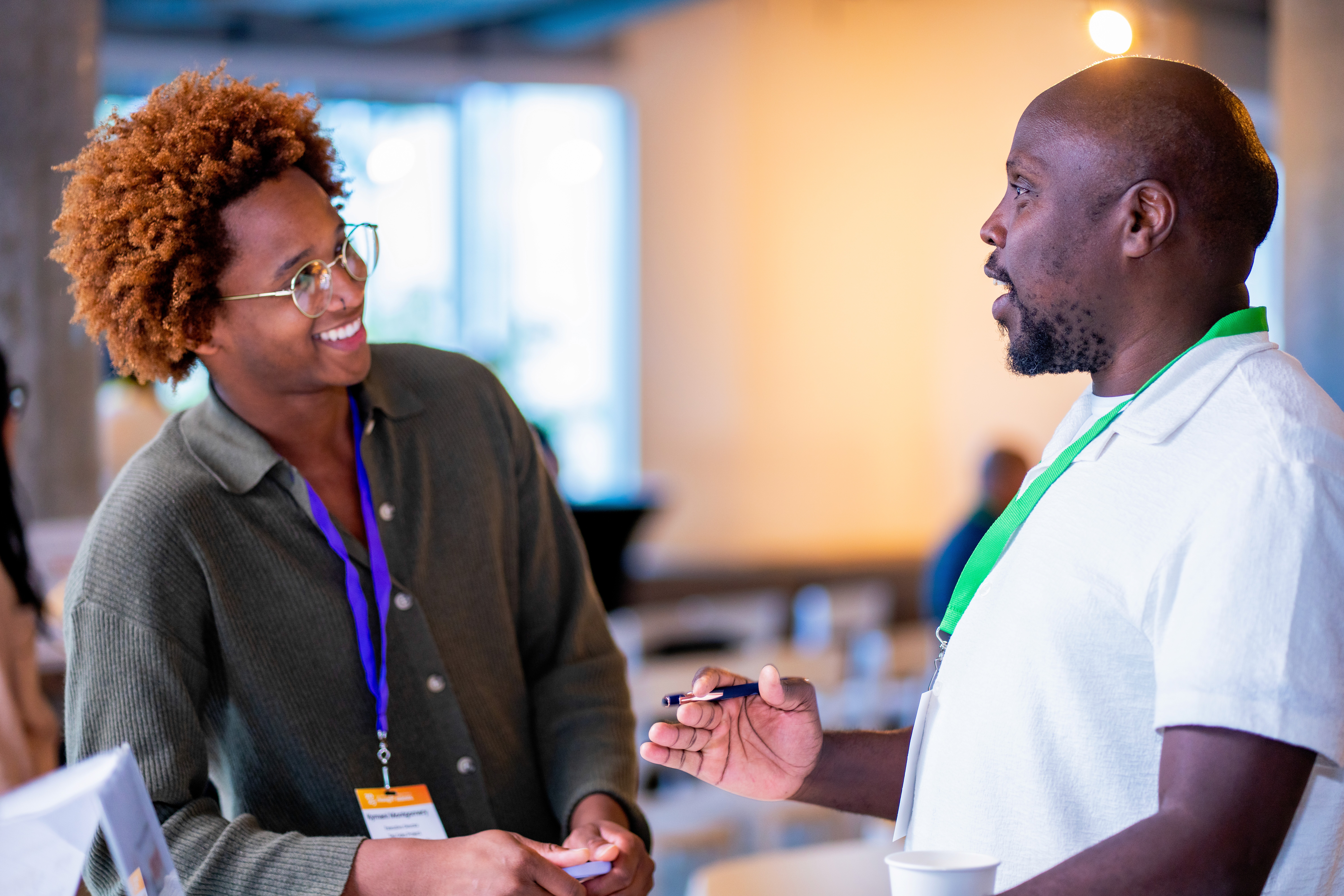 Two people engaged in a discussion, smiling, in an informal indoor setting with bright lighting.