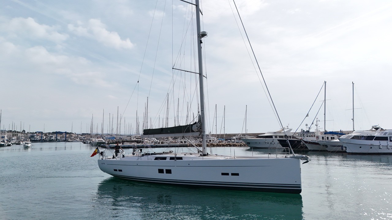 Beneteau Clipper 361 sailing yacht at anchor in Kalkara Creek, Valletta Harbour, Malta, showing the starboard profile with clean white hull, tall aluminium mast, furled sails, bimini and solar panel, with superyachts and the historic Corradino waterfront fortifications visible in the background under a bright summer sky