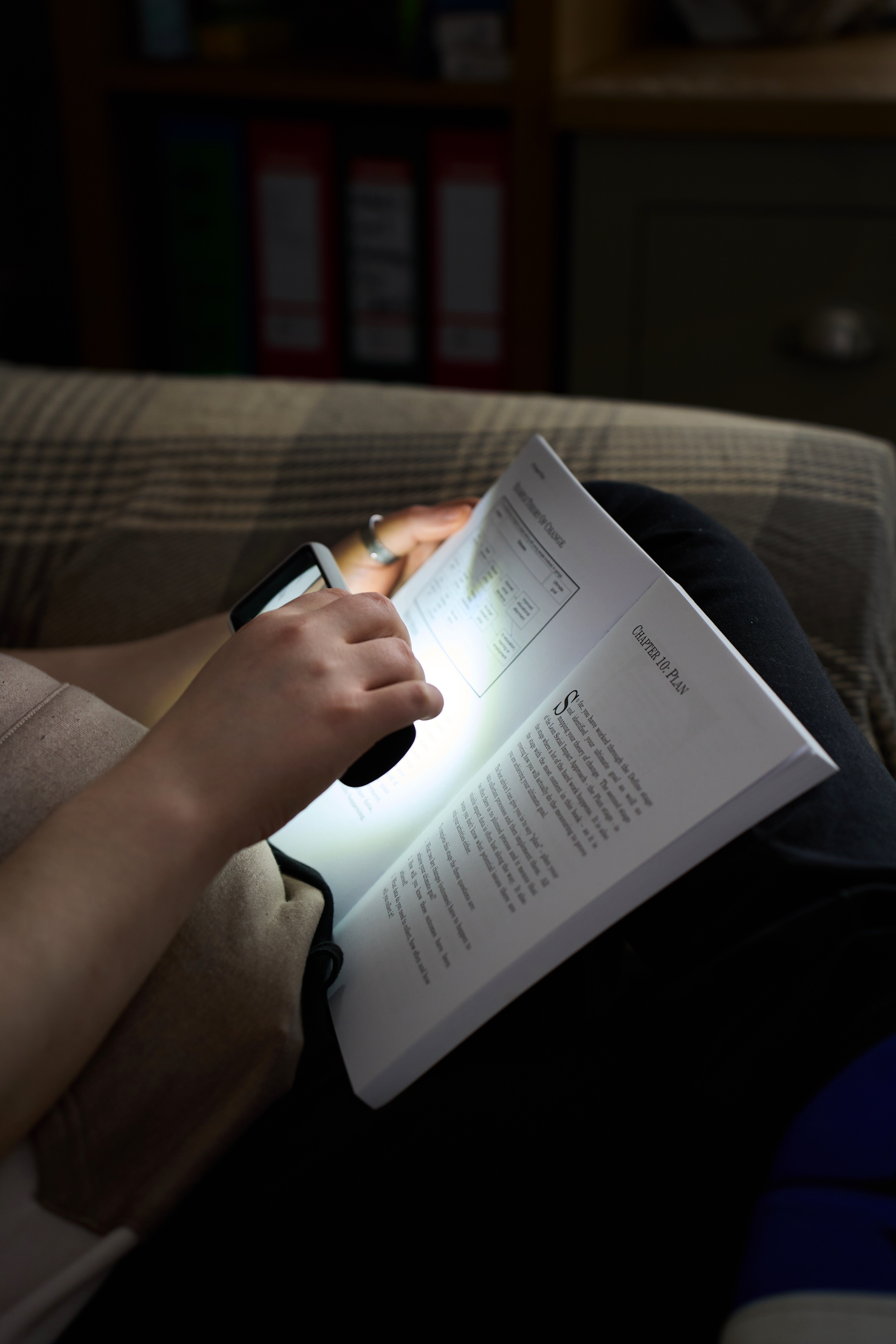 An image of a man using a light and magnifying tool to read a book, whilst sat on a couch.